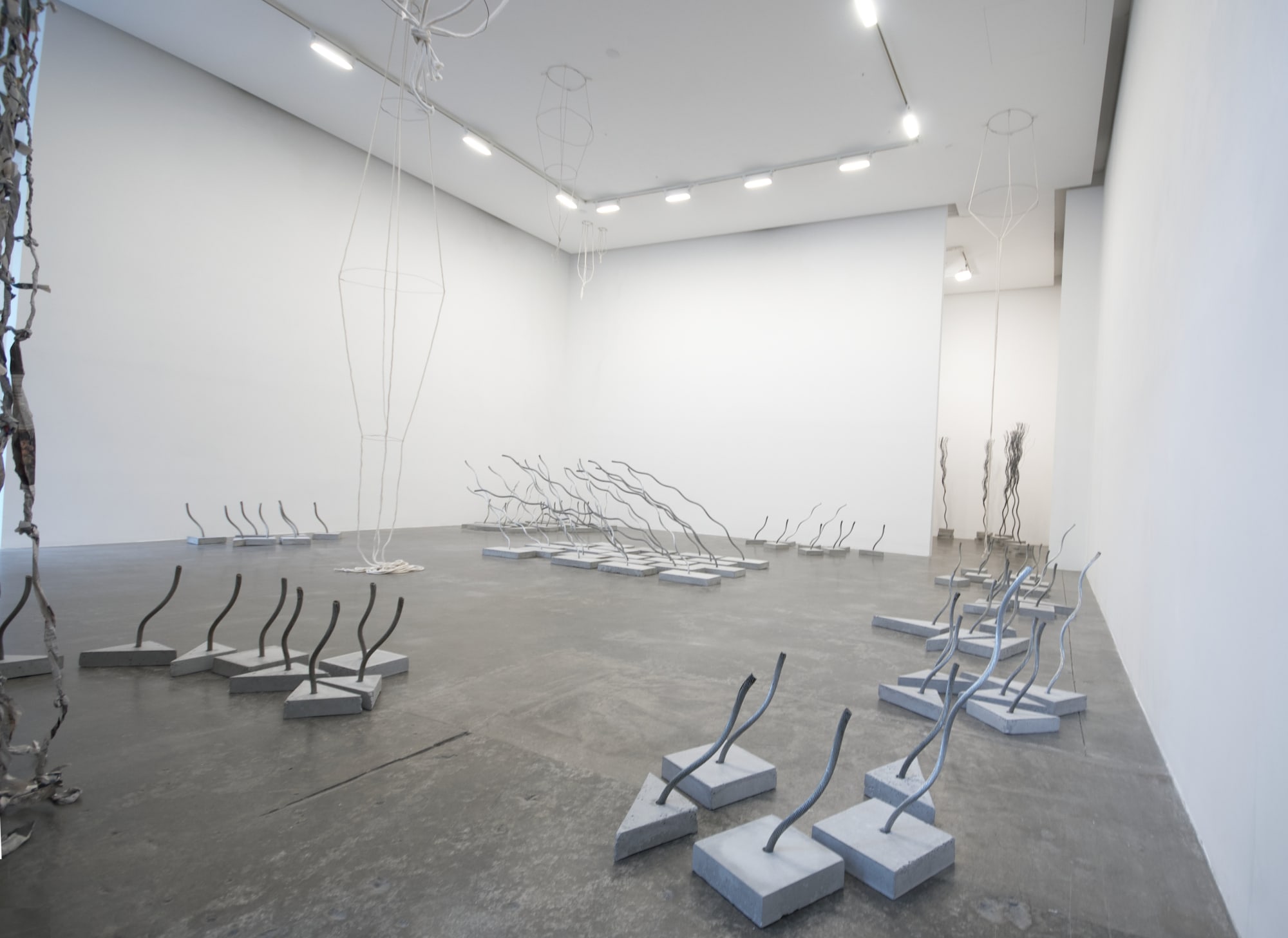 An installation view showing several arrangements of sculptures made of wire rope embedded into concrete bases as well as a curtain of twisted newspaper and a rope and wire sculpture hanging from the ceiling 