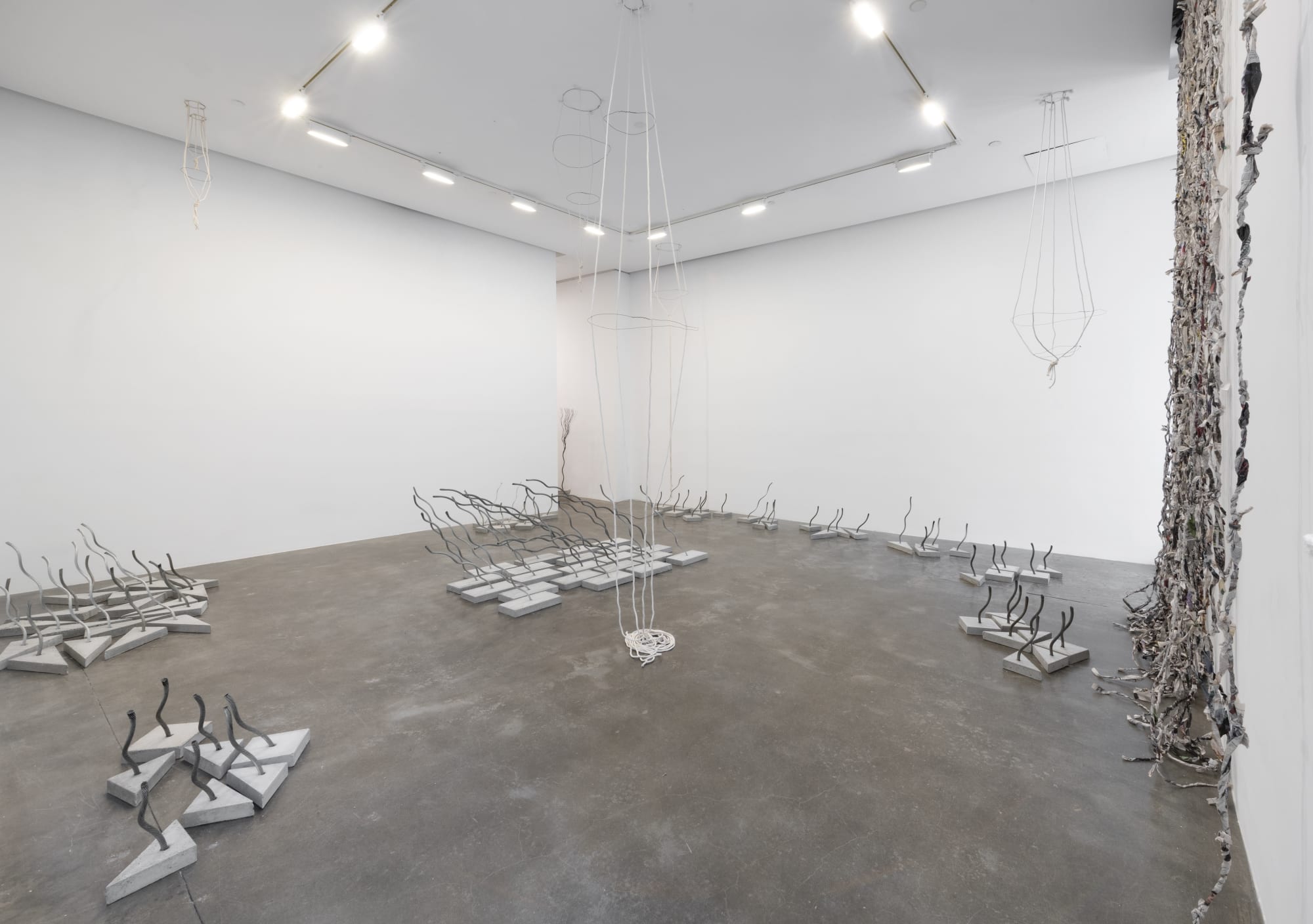 An installation view showing several arrangements of sculptures made of wire rope embedded into concrete bases as well as a curtain of twisted newspaper and a rope and wire sculpture hanging from the ceiling 