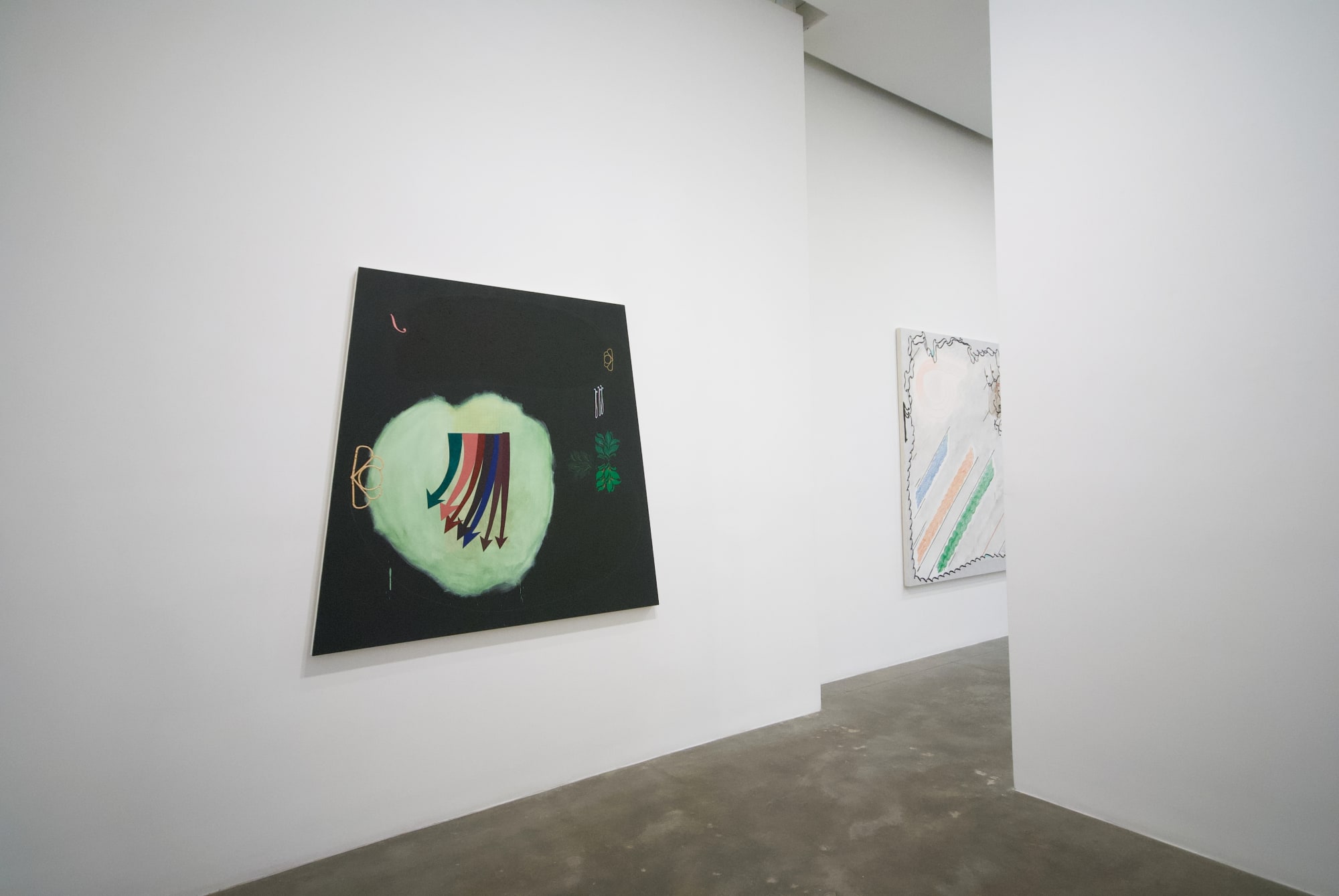 Installation view taken at a side angle of a black trapezoidal canvas with a large, pale green circle off center and multicolored arrows pointing downward, as well as symbols and leaves scattered throughout the composition