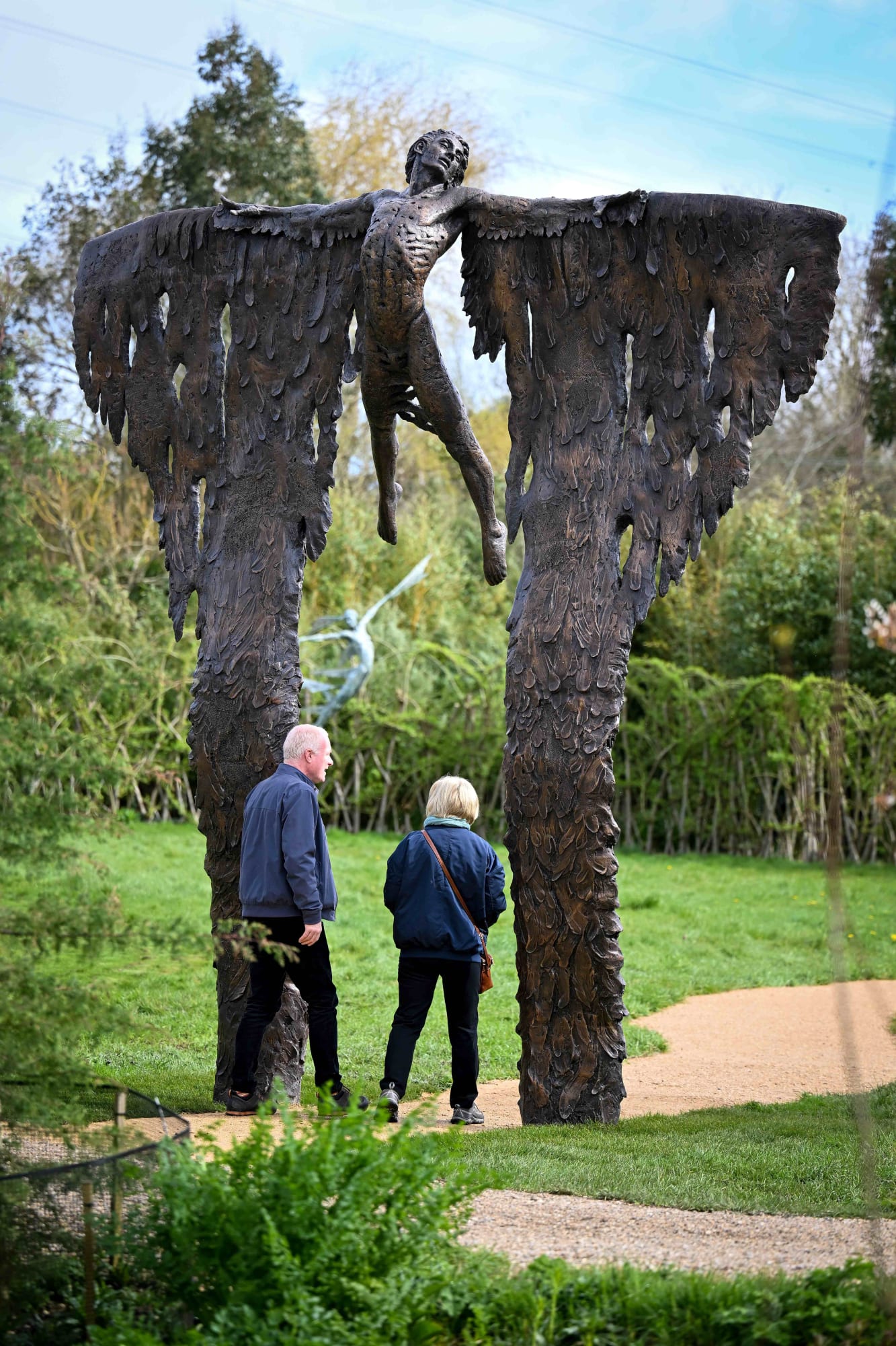 Two people are walking between the wings of the monumental bronze sculpture by Nicola Godden of a mythological figure at a Dorset sculpture park.