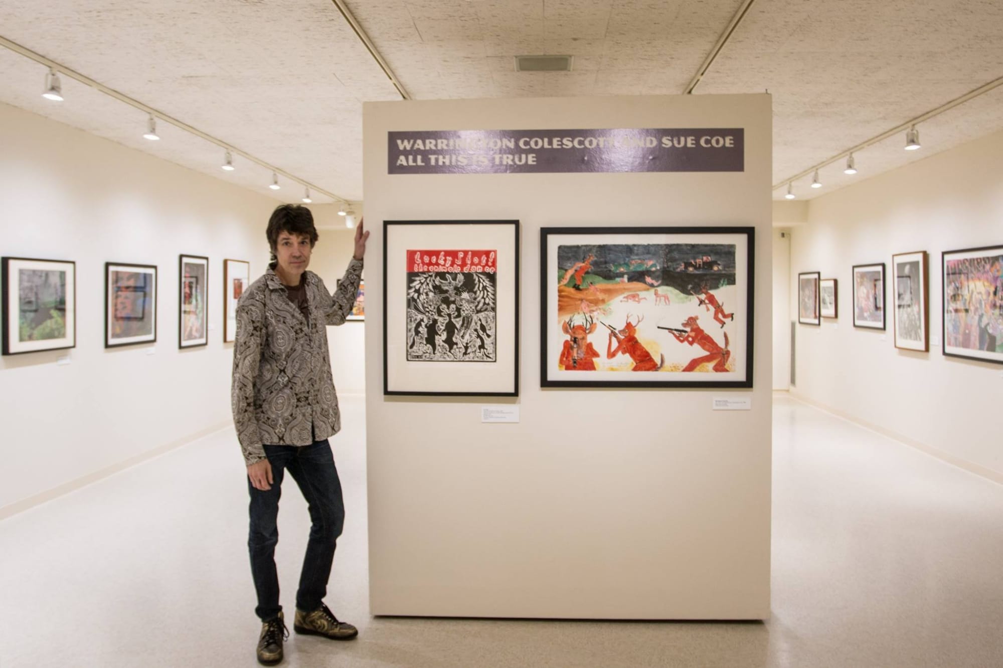 Photograph of John Schuerman, the curator of the exhibition, standing next to the entrance of the exhibition "All This is True." The wall behind him has one piece of art by Sue Coe and one piece by Warrington Colescott.