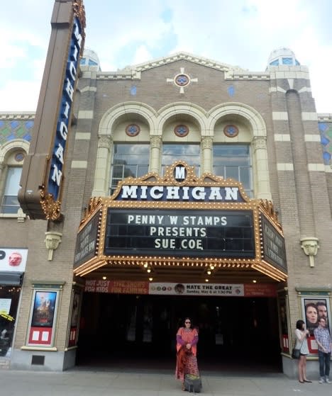 The artist stands in front of a theater. The theater's billboard announces that Sue Coe will be giving the lecture for the Penny Stamps Distinguished Speaker Series