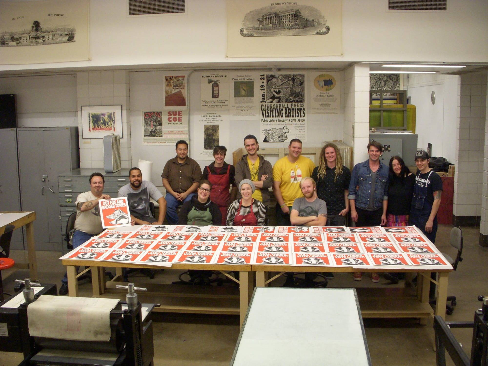 The artist stands with a group of professors and students at the university's print workshop. They stand behind a table full lithographs left out to dry.
