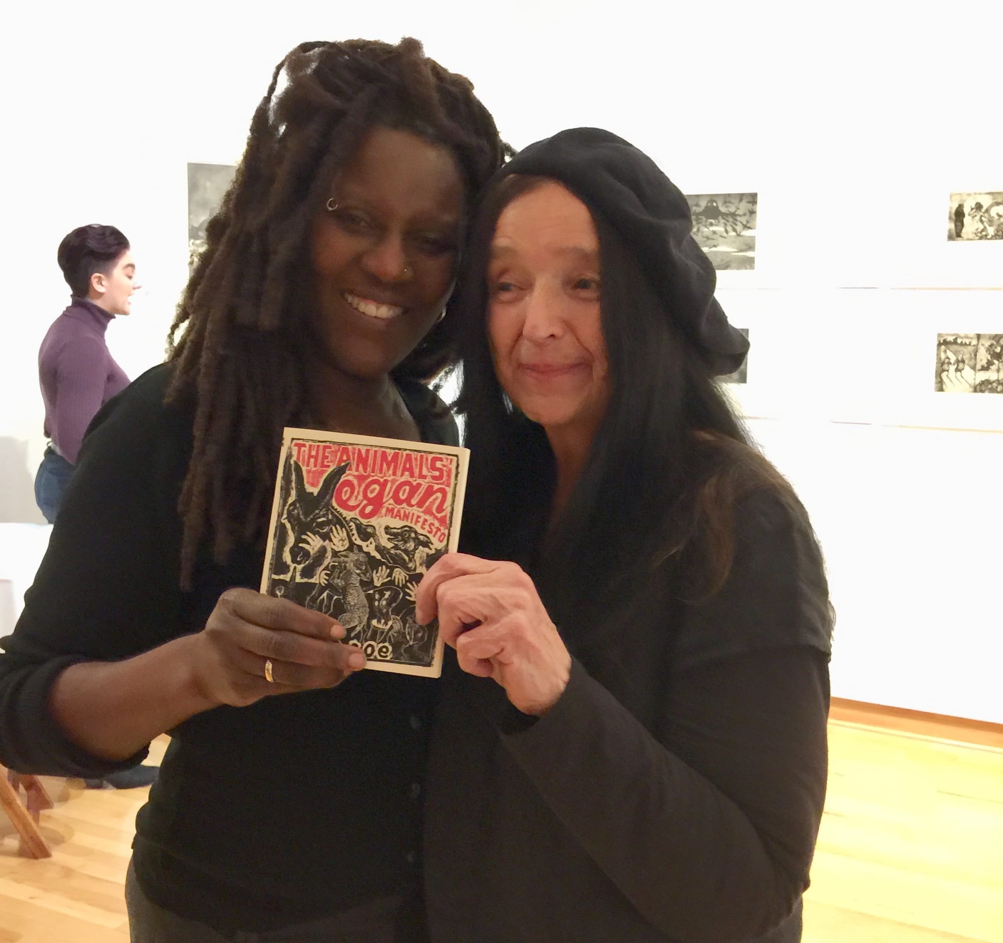 The artist and a professor stand together in an art gallery holding up Coe's book "The Animals' Vegan Manifesto"