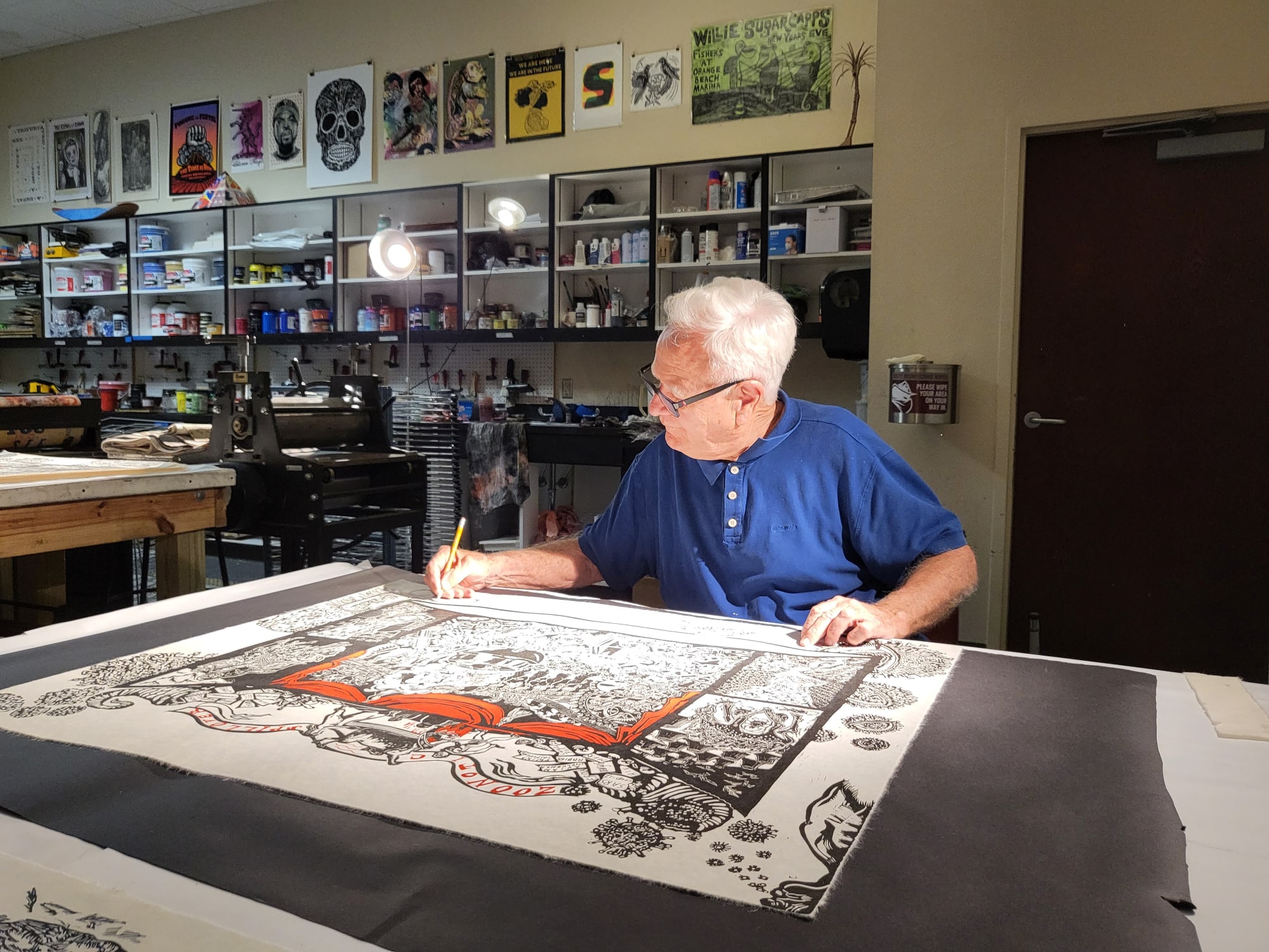 Artist Dr. Eric Avery wears a blue shirt and sits to sign impressions of his collaboration with Sue Coe "Zoonotic Spillover" 2023
