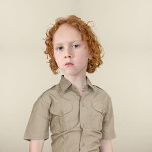 Half-length portrait of a boy with red curly hair, wearing a short-sleeved khaki military-style shirt, gazing at the viewer.