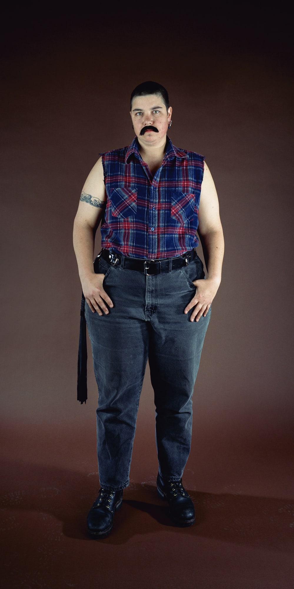 Full length standing figure with a tattoo around her right bicep, wearing a sleeveless plaid shirt, blue jeans, and a fake mustache, against a plain brown background.
