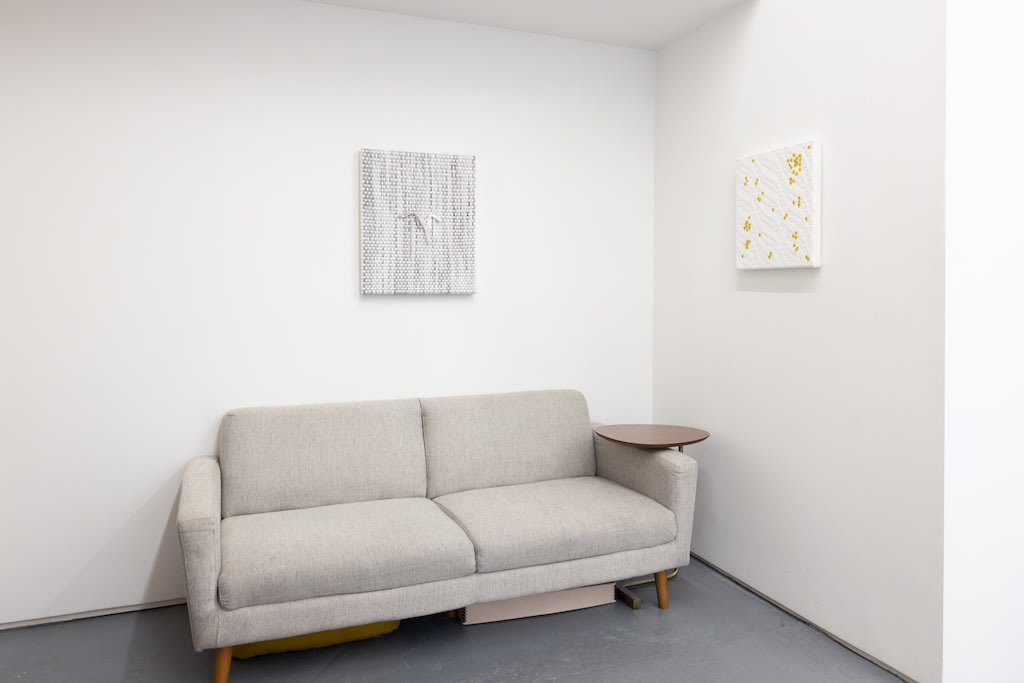 Installation of Didi Rojas exhibition - two shoelace woven paintings on wall (painting on right has yellow smiley face charms scattered on surface) with a couch in center