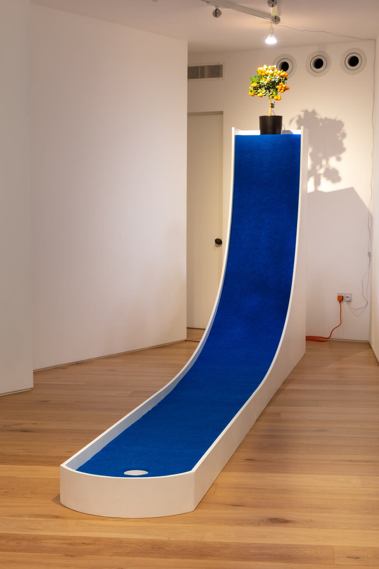 Clara Hastrup Fruitplayer (hole 1), 2023 Calamondin citrus tree, customised wooden mini golf ramp, blue putting carpet, putting hole, putting flag, grow light, vibration motor, Arduino, plastic safety storage boxes, electrical wires, customised metal support structure, wind sensor