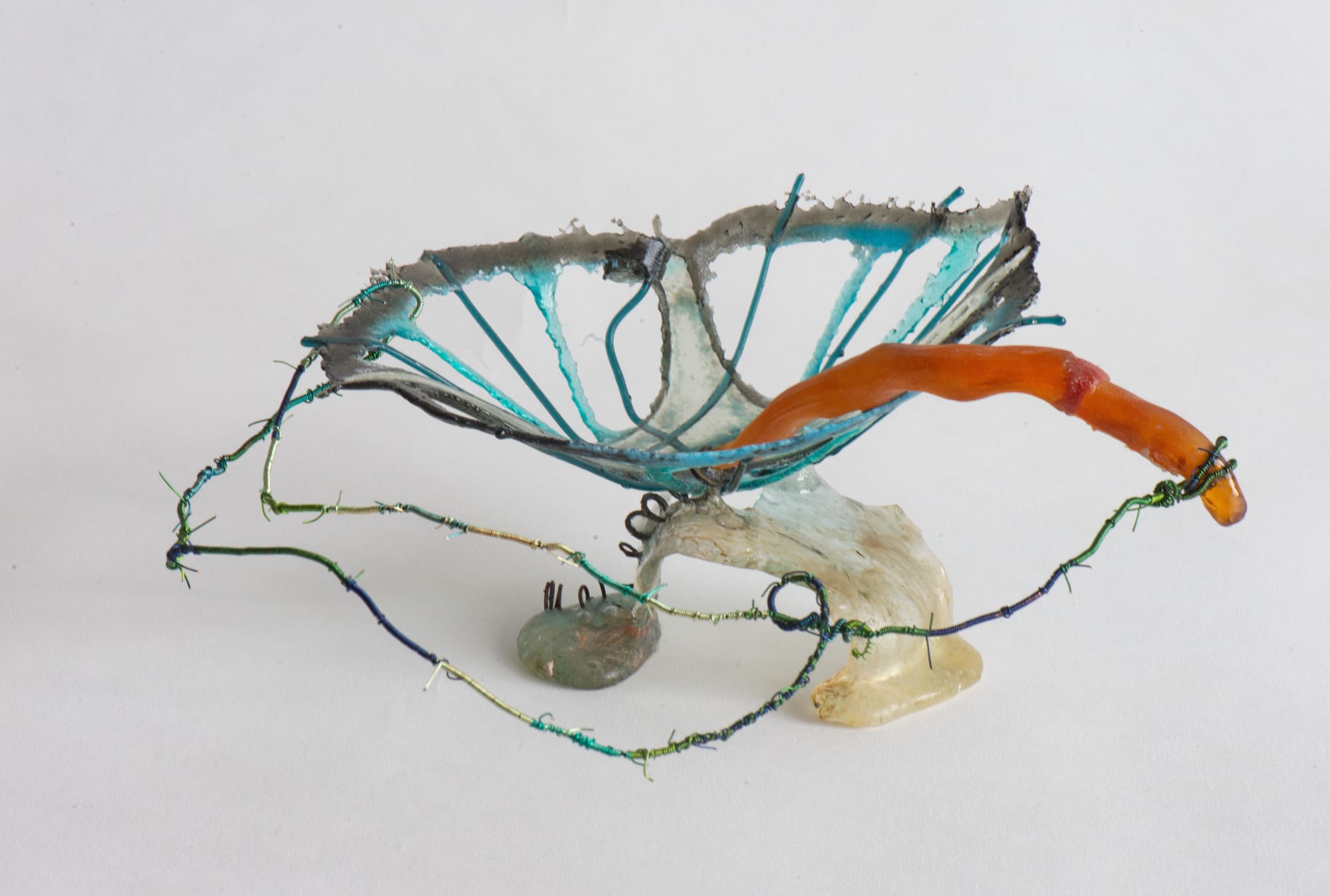 Nancy Cohen's "Momentary" art piece, made from glass, metal and wire in shades. The work has shades of blue, orange, and white. The blue metal object resembles a flower while the orange glass string looks like a worm crawling out of the flower.