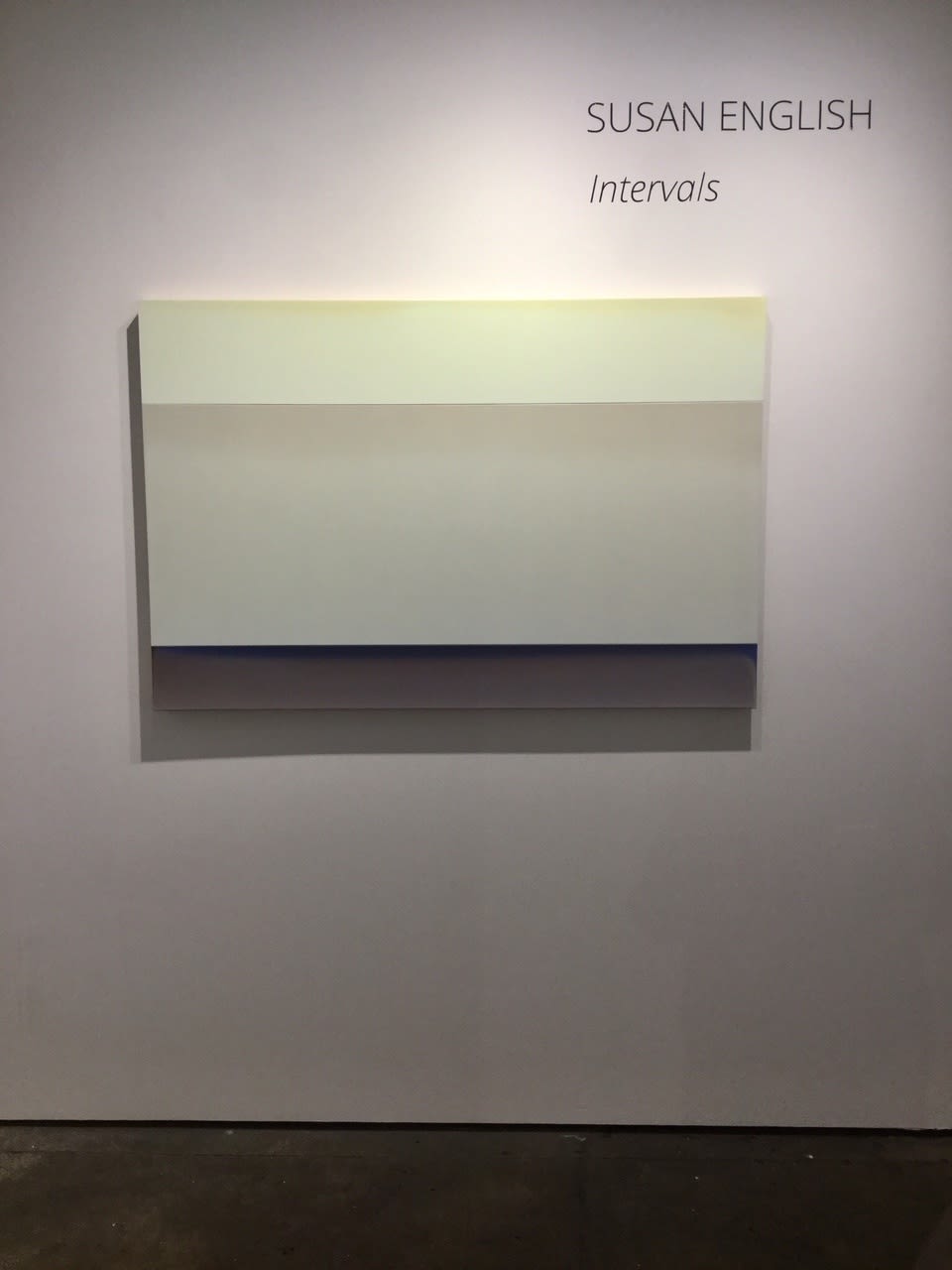 Installation shot of Susan English's "Lift", a tinted polymer on panel in shades of tan, cream, blue and gray. The work sits under wall lettering reading, "SUSAN ENGLISH - Interval".