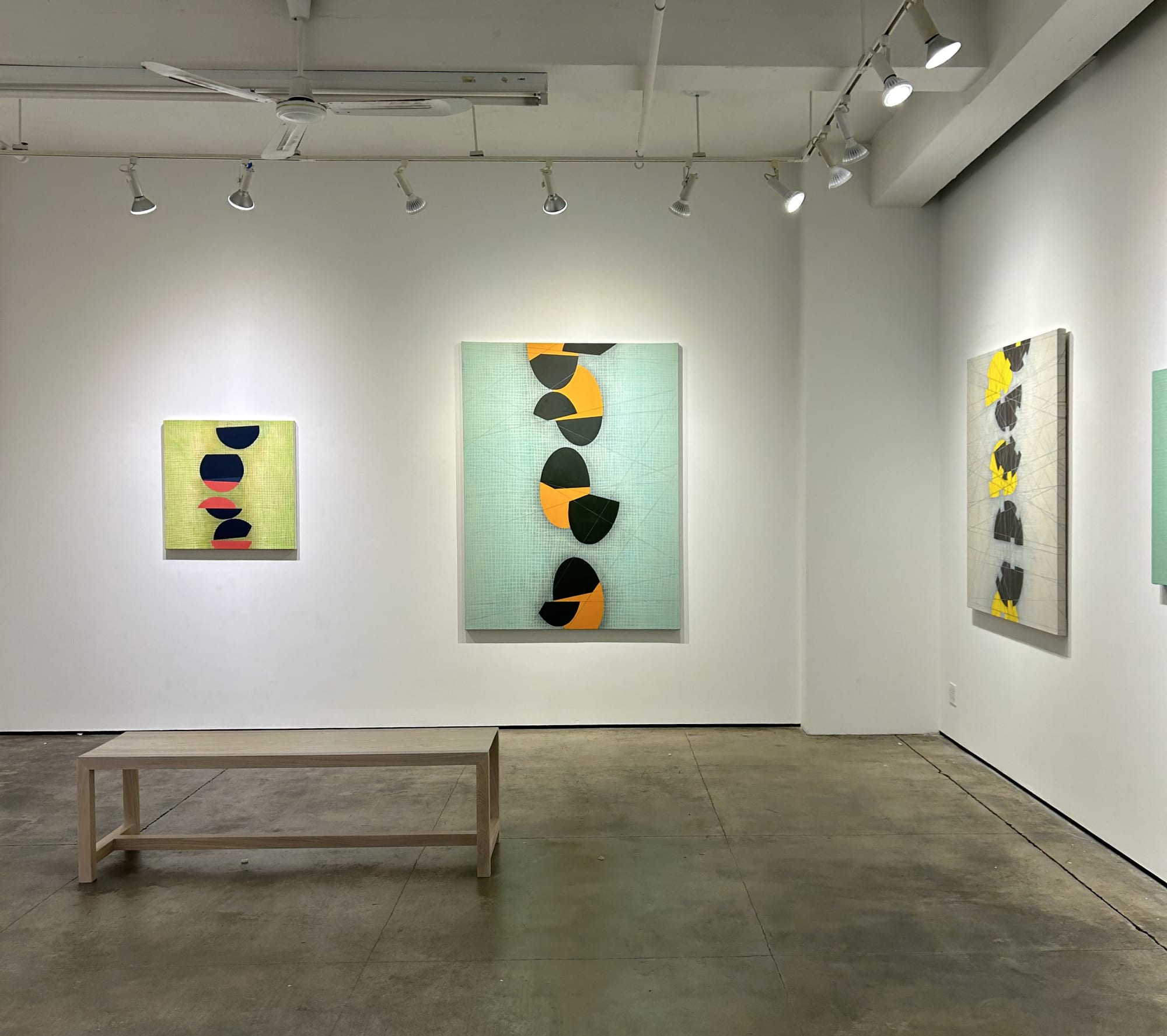 Photo of a painting exhibition, with three works visible, and a bench in the center of the room.  Each painting has a low contrast painted grid in the background, with a series of circular shapes going up the center, which appear to be bisected at various