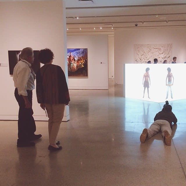 A man lies on the ground photographing an artwork in the distance as a couple observe the same work.