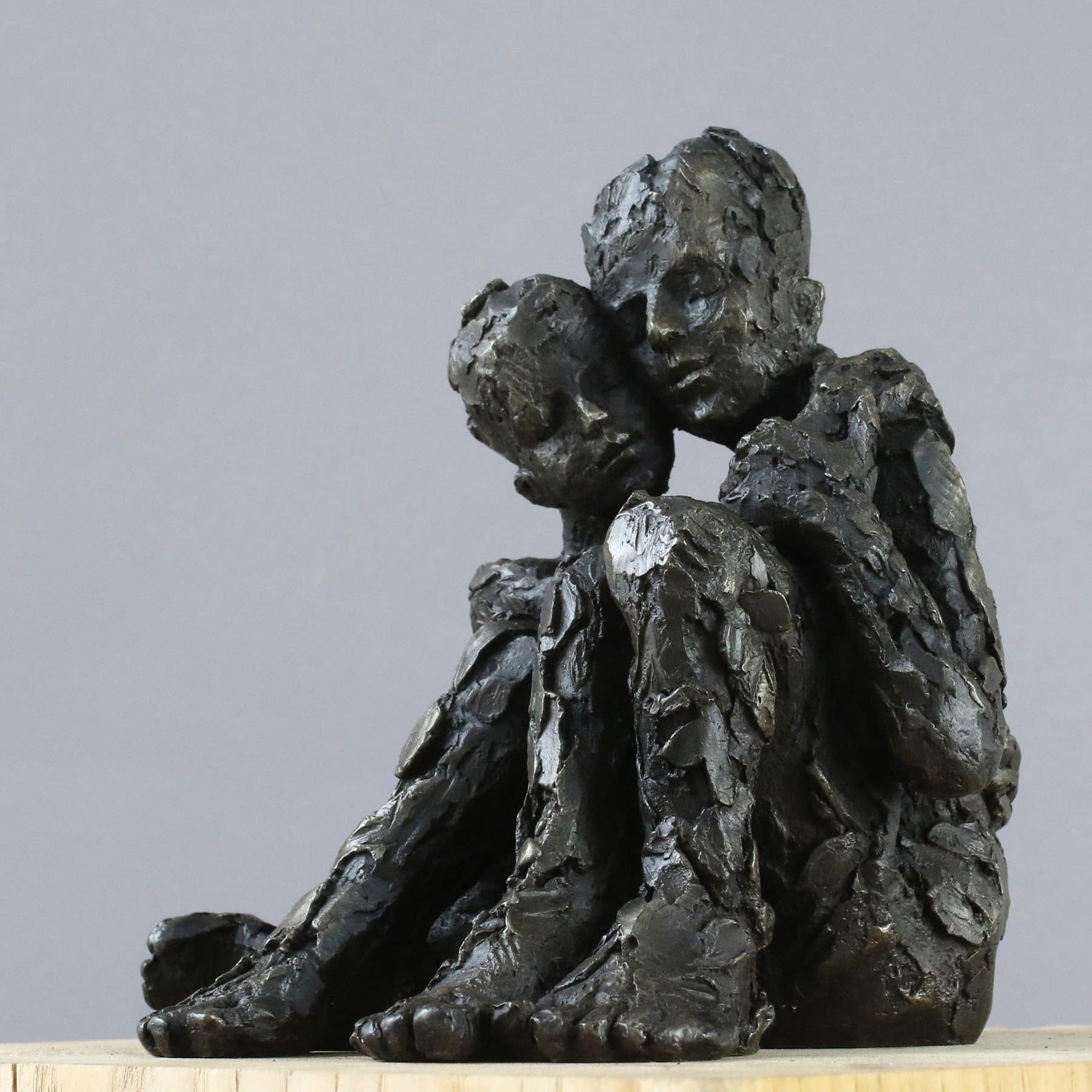 Carol Peace sculpture of a couple, sat side by side, embracing, with their heads close together. The surface is a rough texture and the colour is dark almost black.