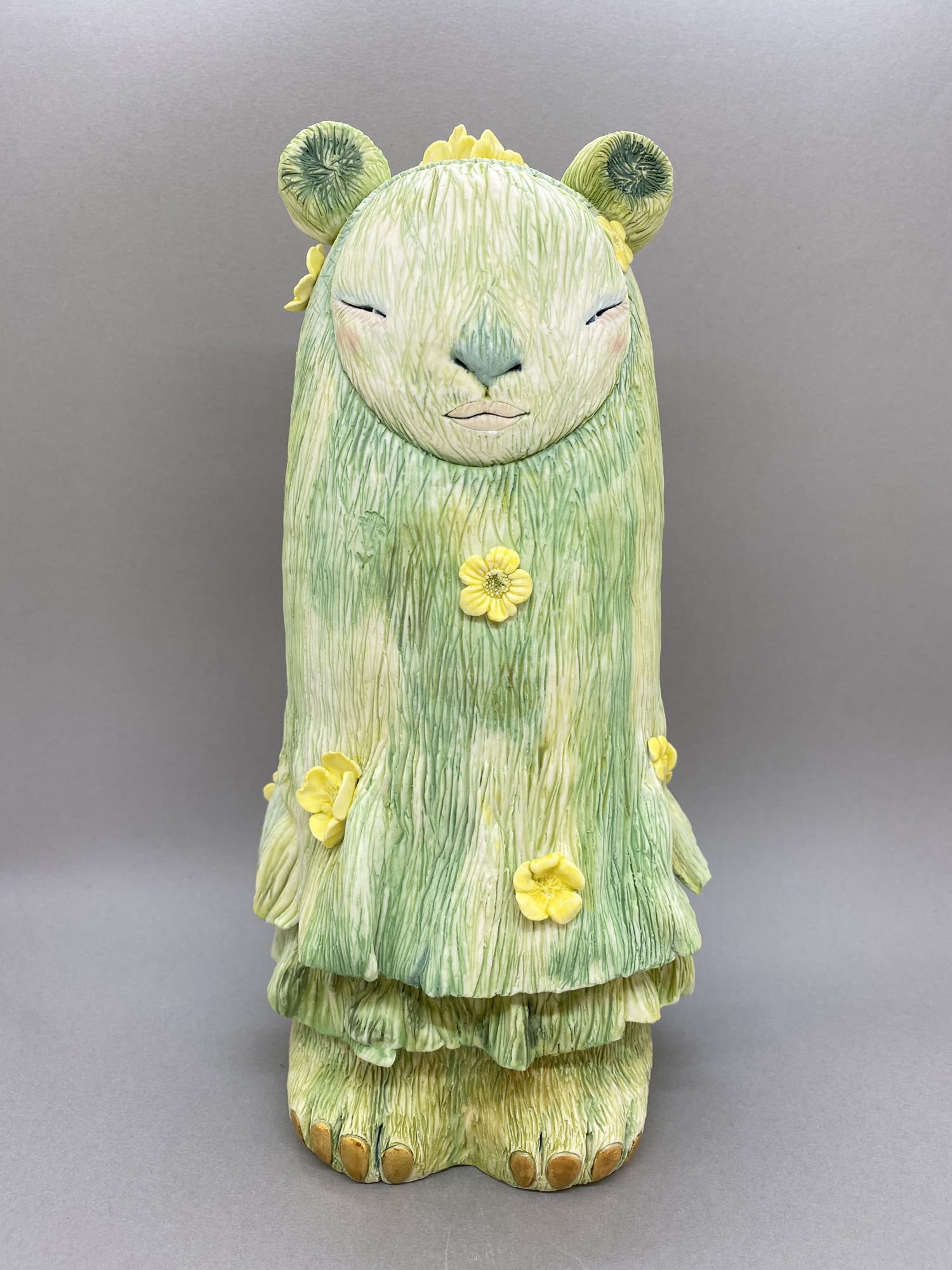 Inspired by Japanese kami, a sculpture in green with grassy detail, small yellow flowers dotted on its body, round small ears on its head.