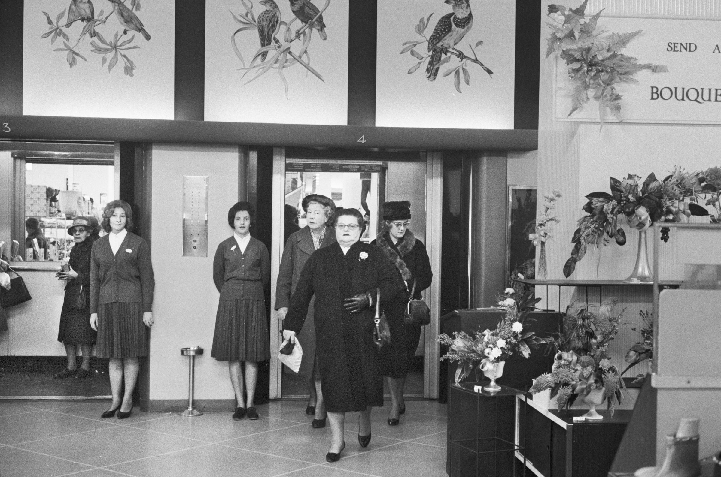 Black and white image of people in full coats and matching pocketbooks and shoes entering a high-end flower store. Store clerks wait by the door.