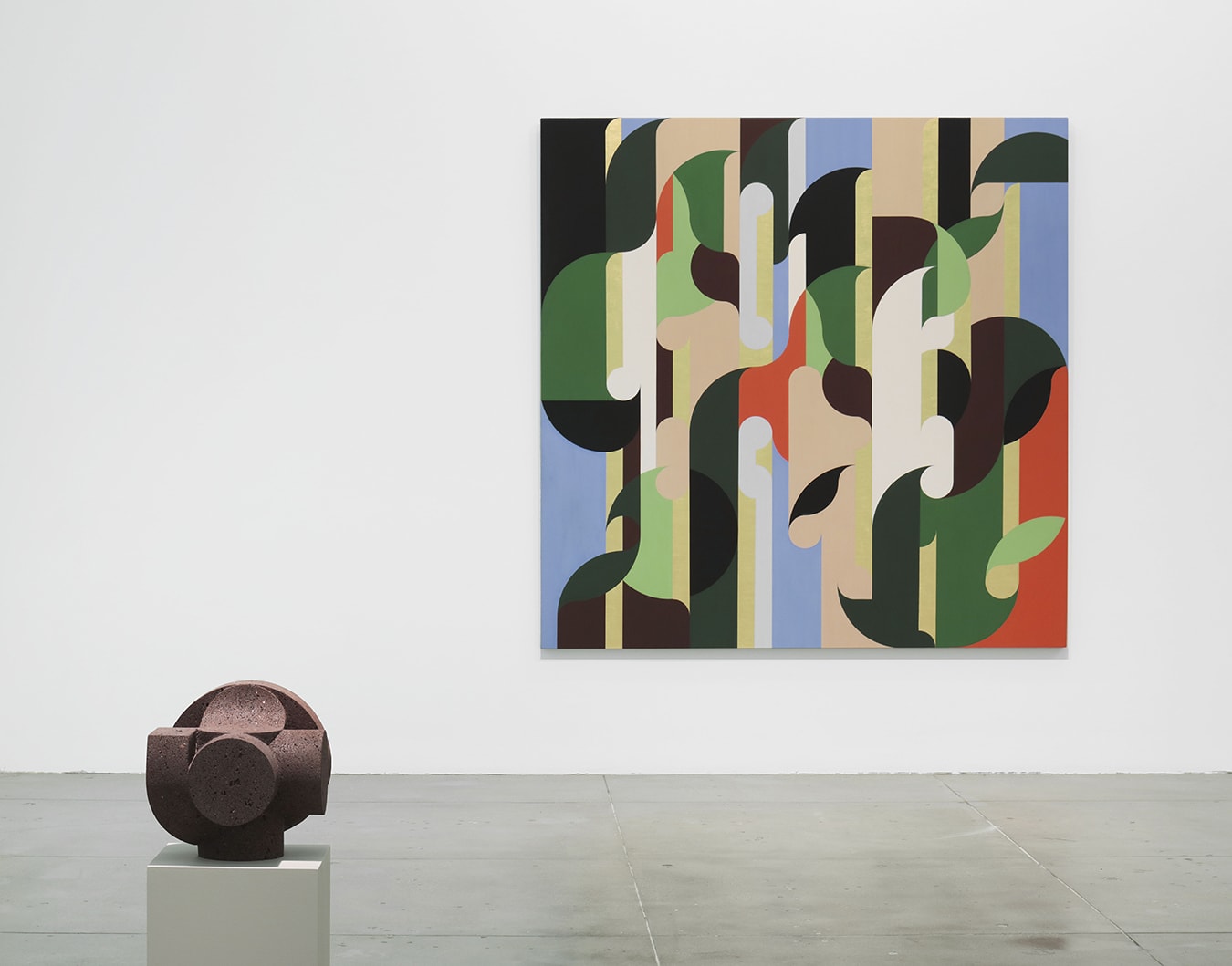 Installation view of a colorful graphically inspired painting on the right side of a white wall with a brown bronze circular sculpture on the floor