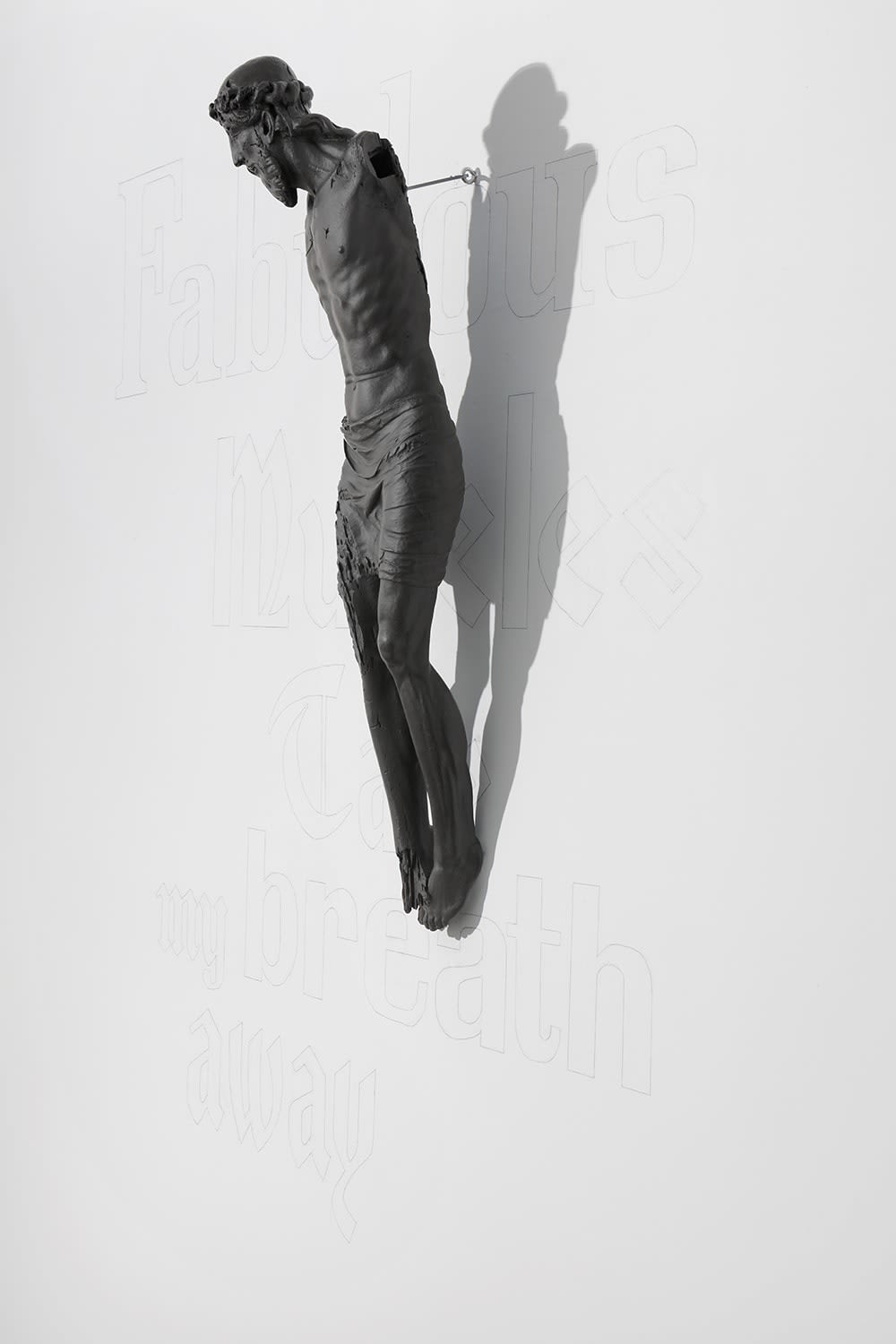 Installation view of a sculpture depicting Jesus crucified, suspended off a gallery wall featuring large faint text.