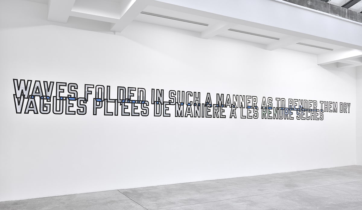 "Waves folded in such a manner as to render them dry" in silver reflective text in English and French on a white wall