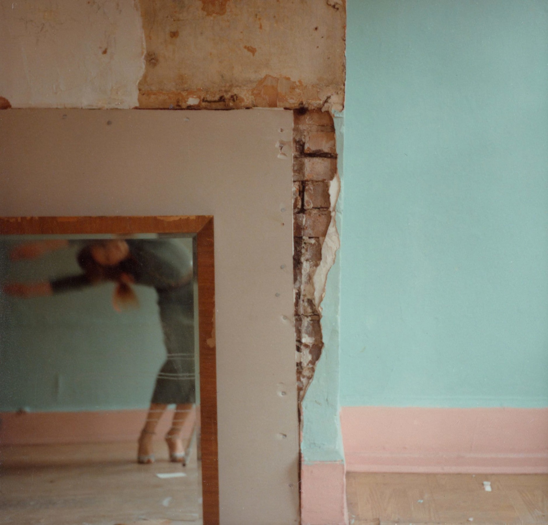 Color photograph of a blue and pink wall with exposed brick and an inlaid mirror, reflecting a blurred bending figure.