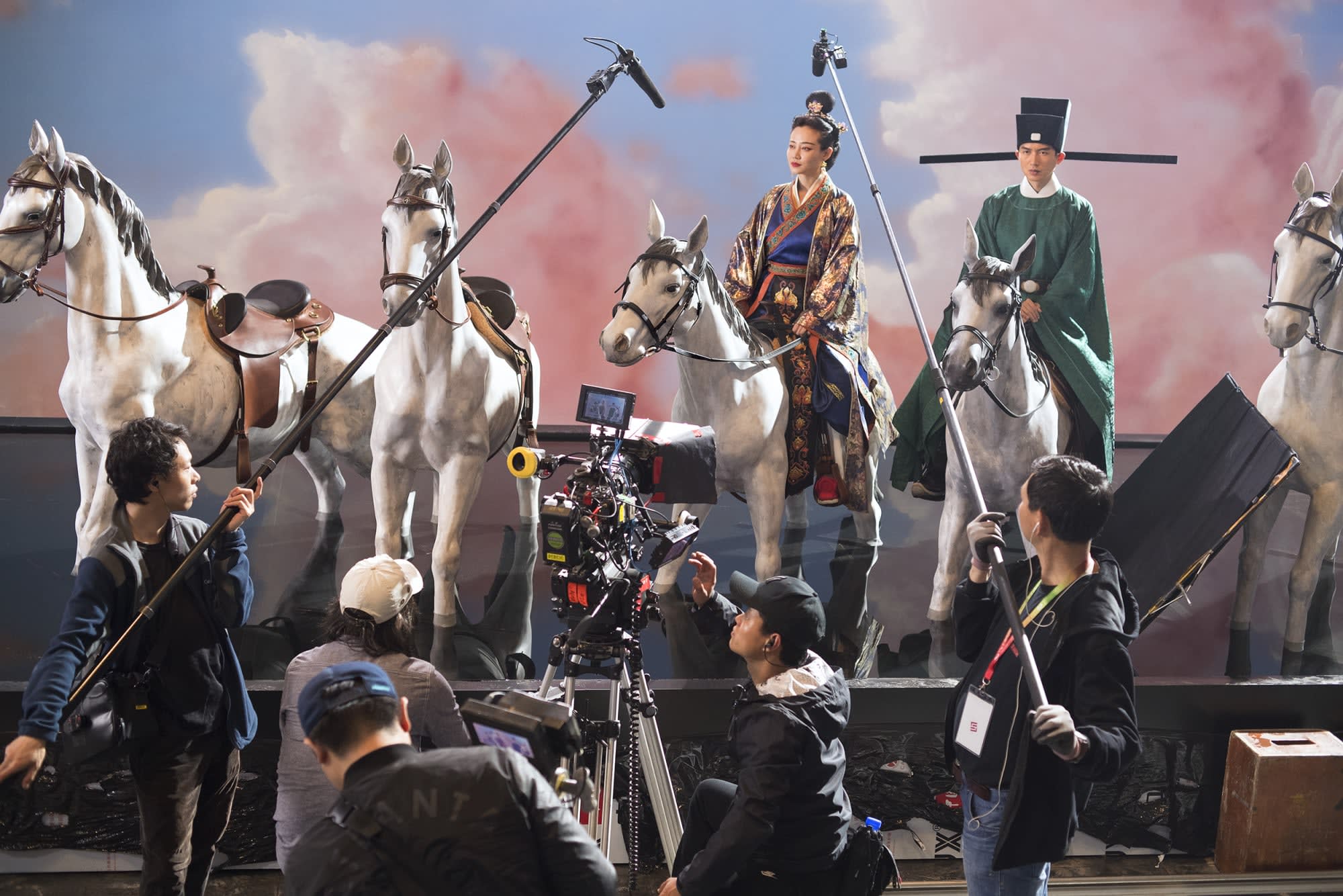 Photograph of five crew members with film equipment documenting a stage set of five horses, two traditionally dressed actors and a backdrop of pink clouds against a blue sky. 