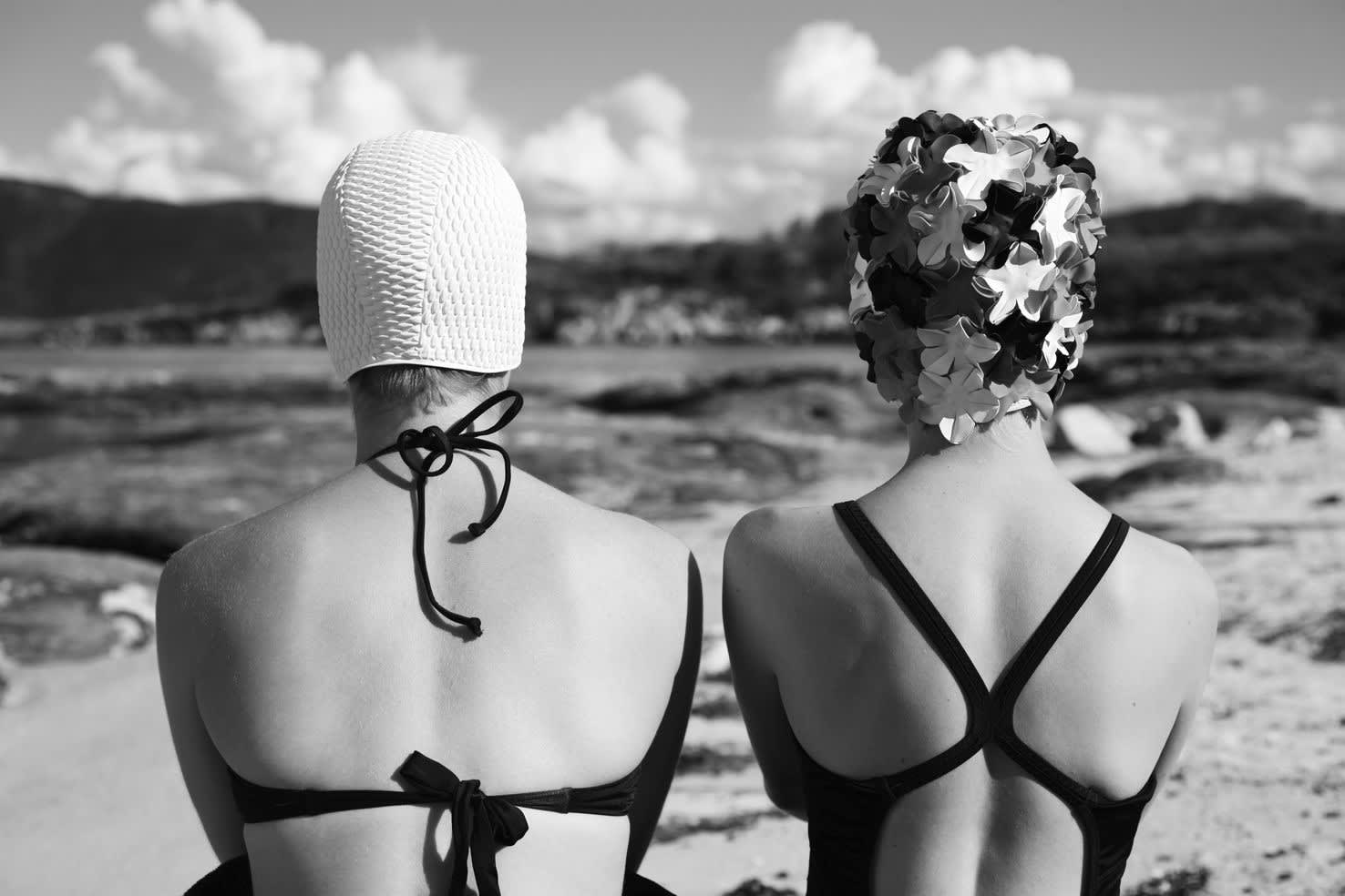 Black and white photograph two figures from behind dressed in swimwear, in front of a blurred landscape.