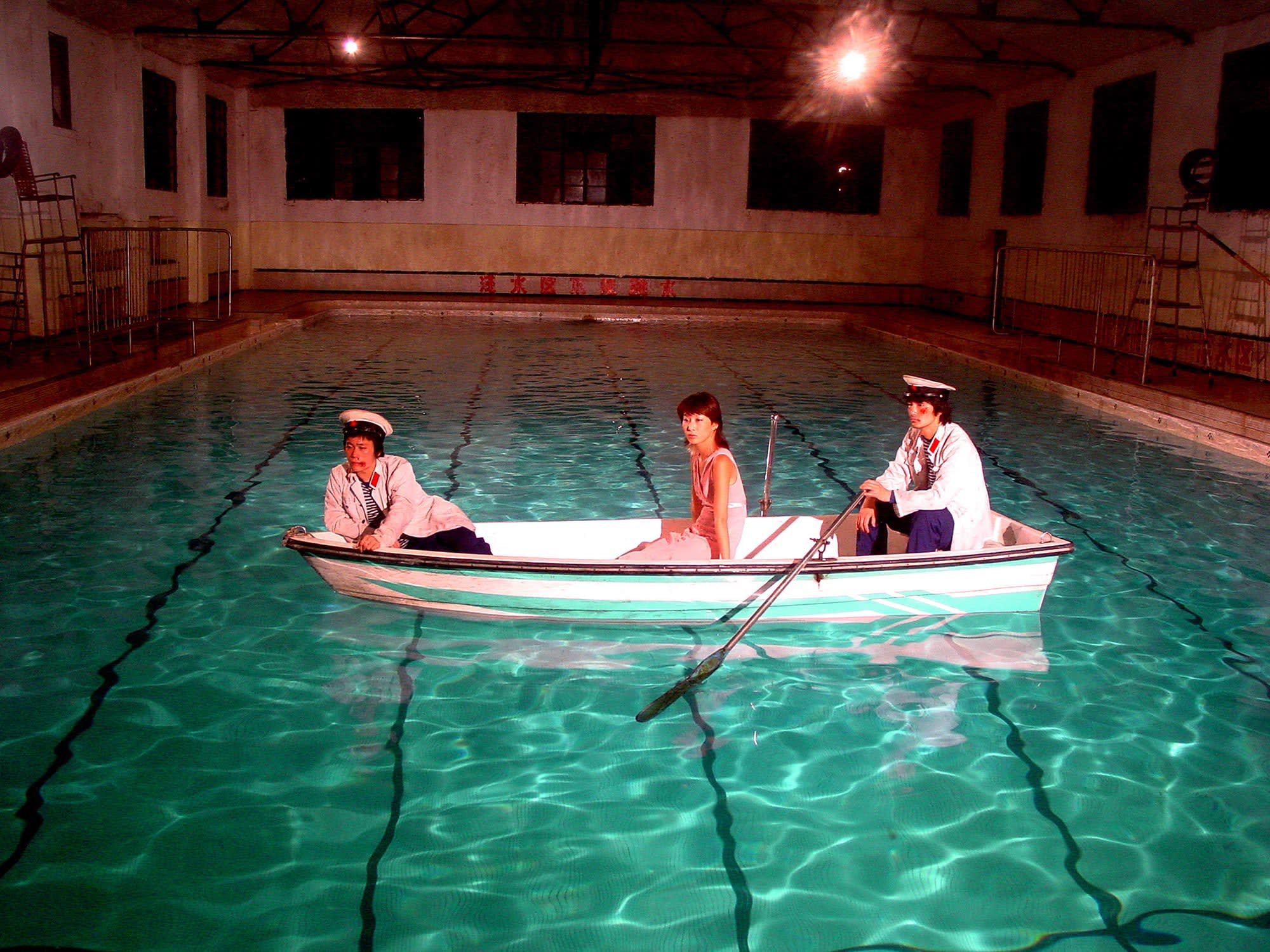 Color photograph of three figures seated in a small boat floating in an indoor swimming pool. 