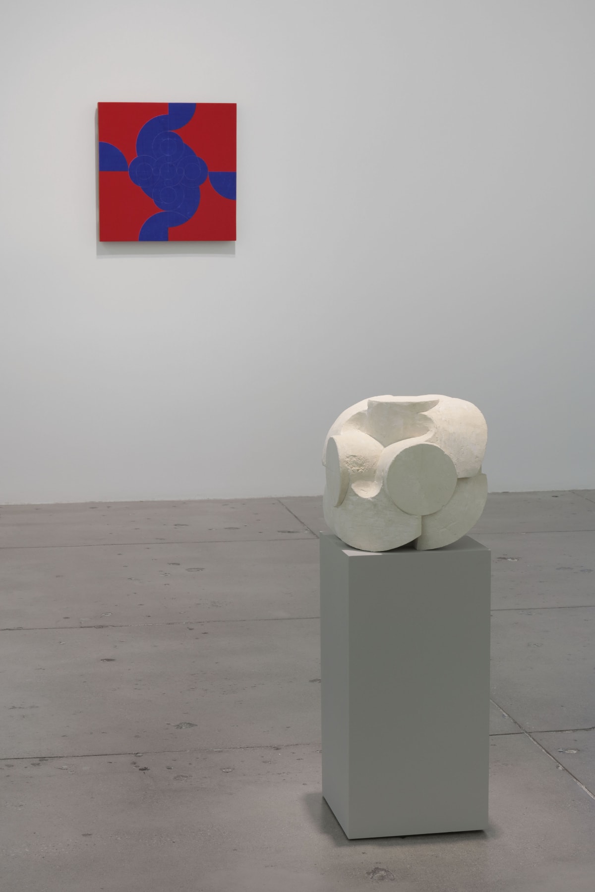 In a large white space, a small red and blue abstract painting hangs on the wall in front of 1 small, white stone sculpture.