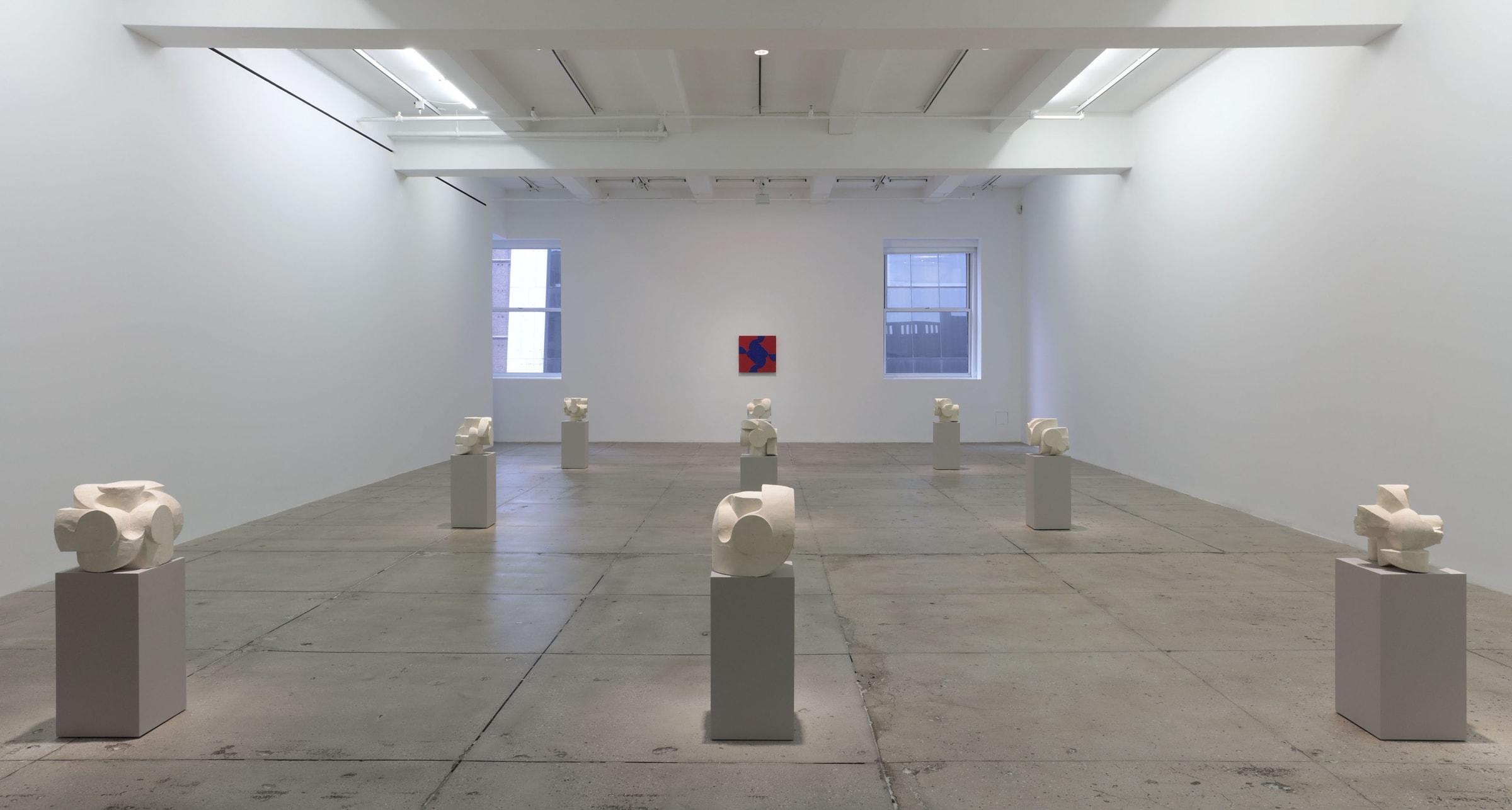 8 small white stone sculptures on pedestals in front of a red abstract painting on a white wall. 