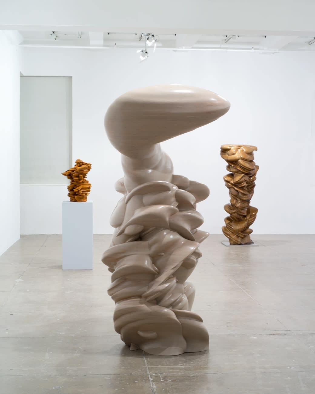 Three vertical abstract sculptures stand in a large white room; the sculpture on the left is on a pedestal. They are beige and brown with both smooth and jagged edges. 