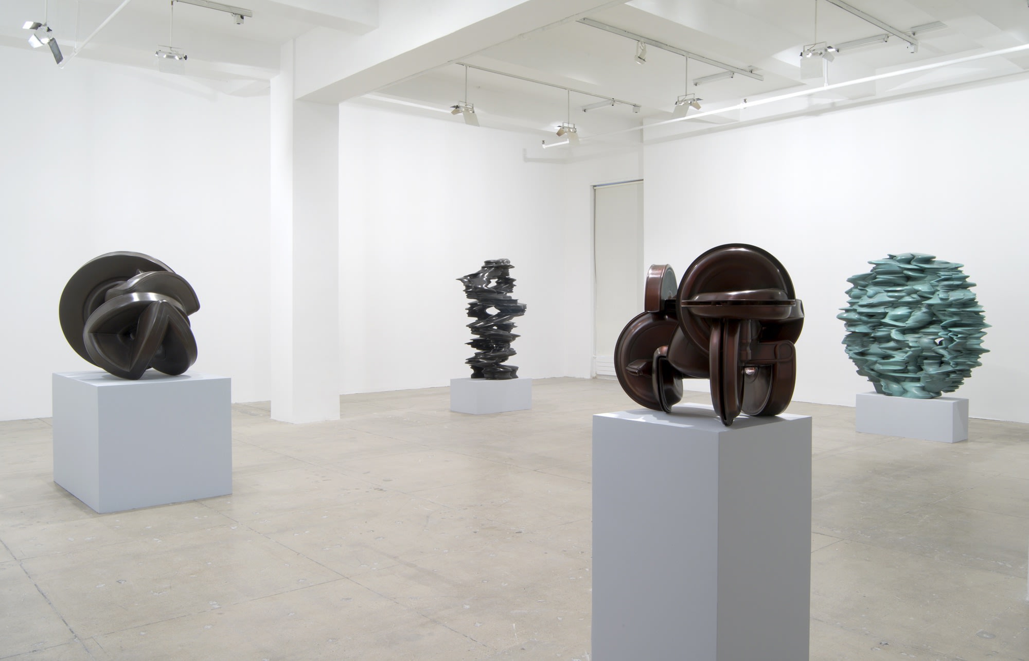 Four abstract sculptures sit on pedestals across a large white room; one on the right is light blue-green and the others are dark brown and grey. 