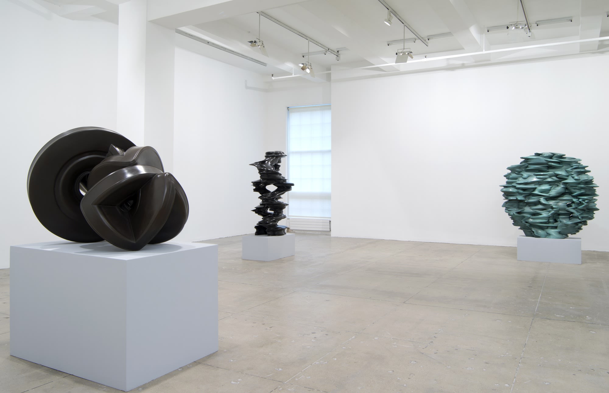 Three abstract sculptures sit on pedestals in a large white room. The one on the right is spherical and light blue-green; the other two are dark grey. 