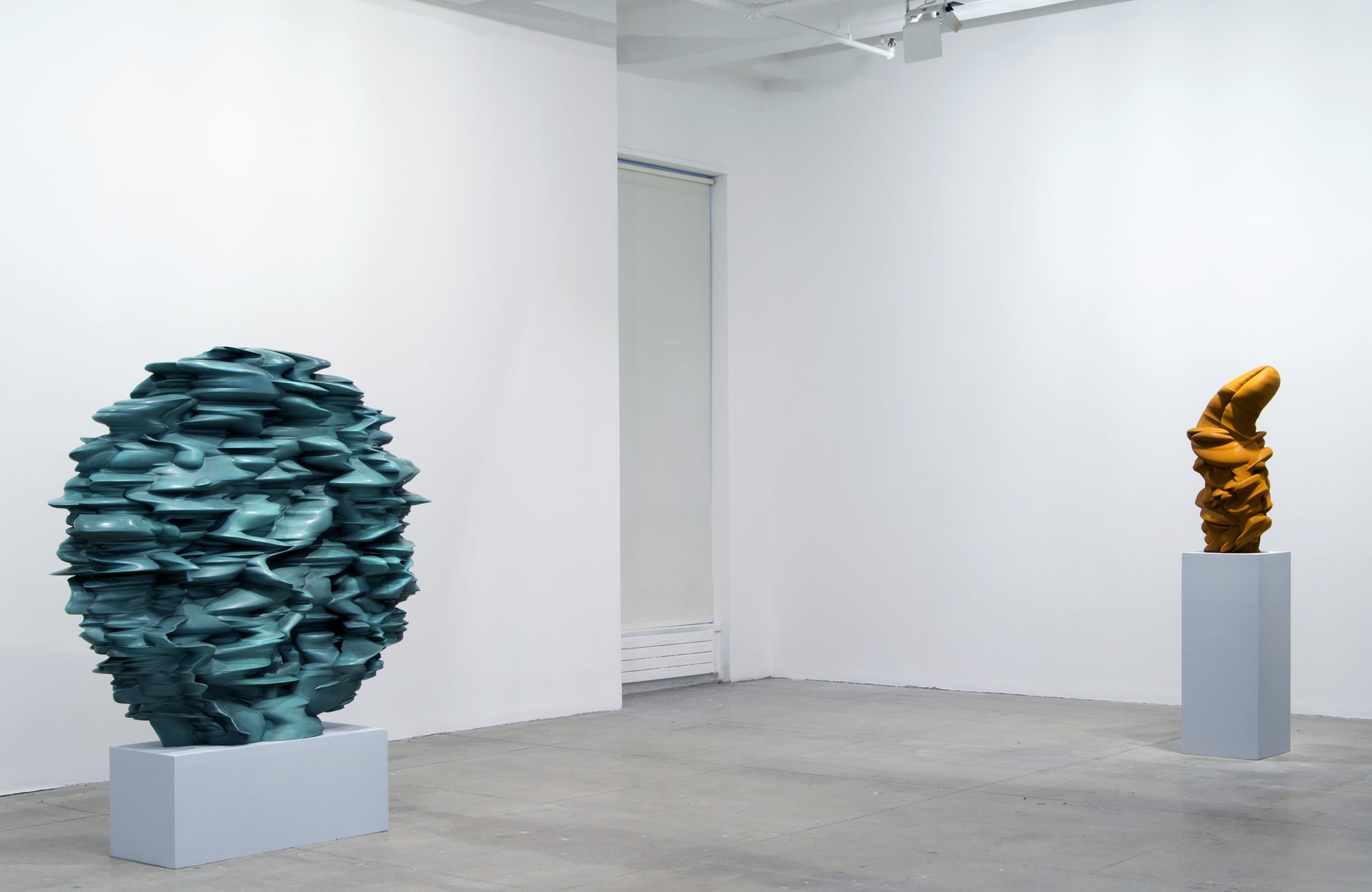 Two abstract sculptures sit on pedestals in a white room. The one on the left is spherical and light blue-green, while on the right the piece is vertical and orange-brown. 