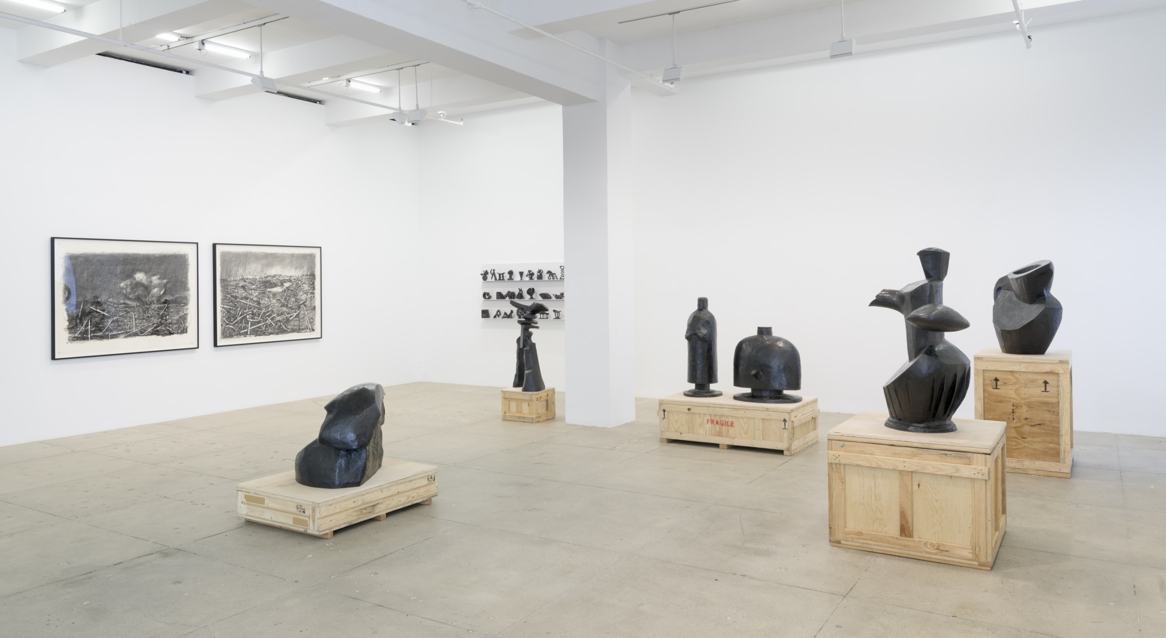 Five large copper sculptures of various figures including vases, men and women, stand next to two dark graphite drawings of war-torn landscapes. 