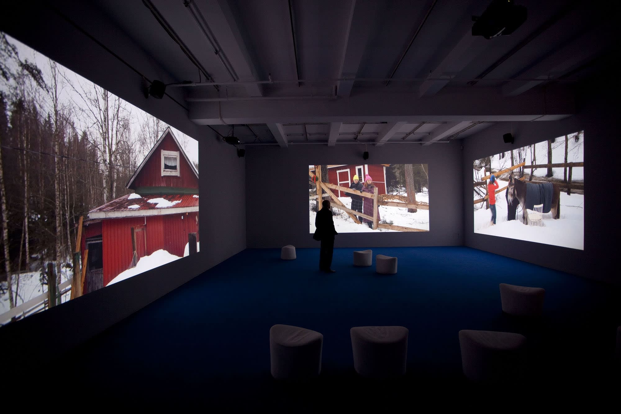 A man stands in the center of a room surrounded by large projections of snow-covered exterior images of a red barn. 