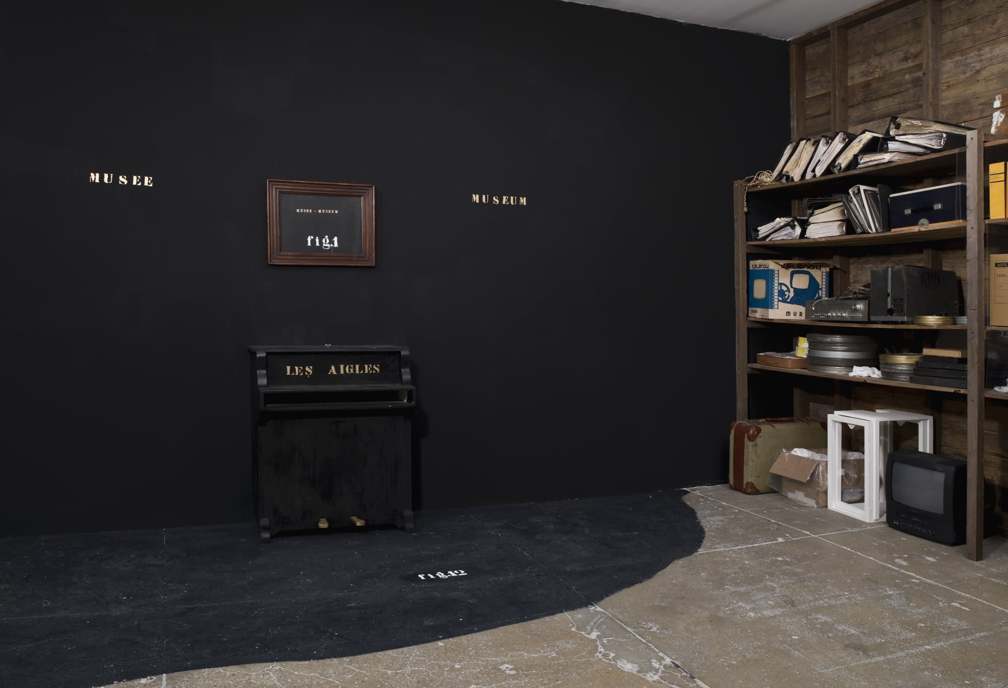 On a black wall, gold text reads MUSEE and MUSEUM on either side of a wood frame containing a black painting that reads the same thing, along with: fig. 1, in white text. Under it, gold text on a black baby piano reads: DES AIGLES. On the right wall, a wo