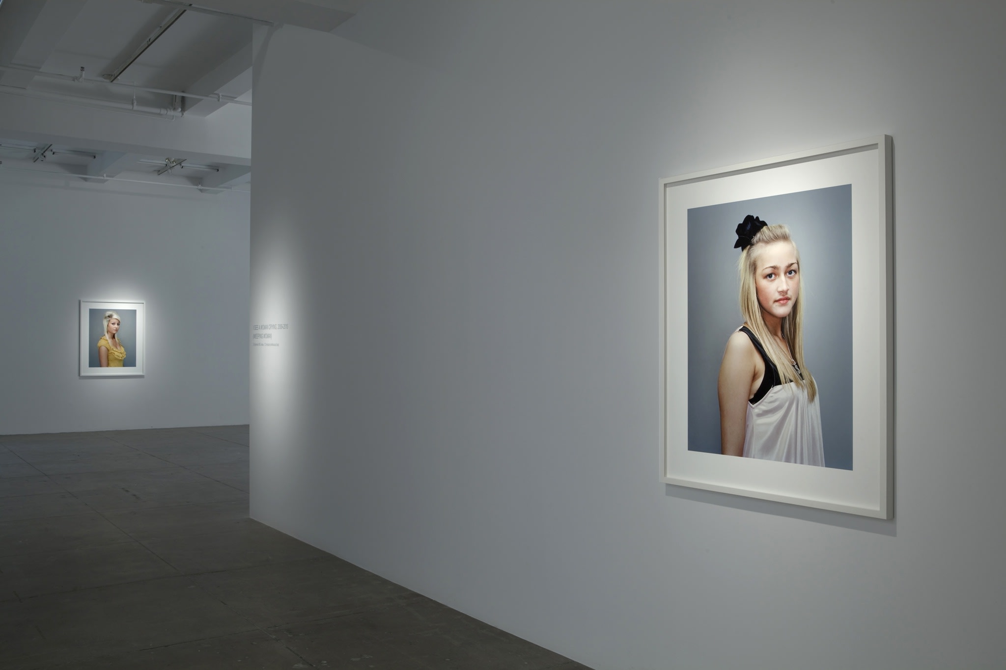 On a white wall, the portrait of a young woman, with blonde hair and blue eyes, wearing a white top with black straps, hangs. In the background, on another wall, a portait shows a woman wearing a yellow top.