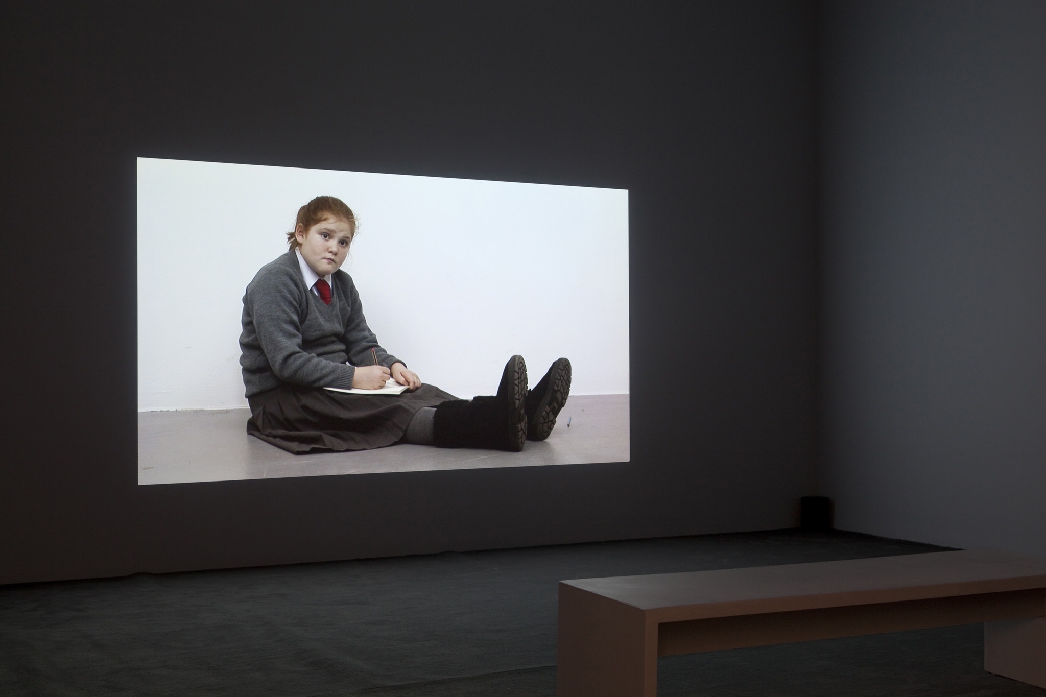 In a dark room, a large projection shows a tween school girl in her uniform (brown skirt, brown sweater over white collared shirt with a red tie, black Ugg boots) sits on the floor, writing in her notebook and staring out towards the viewer.