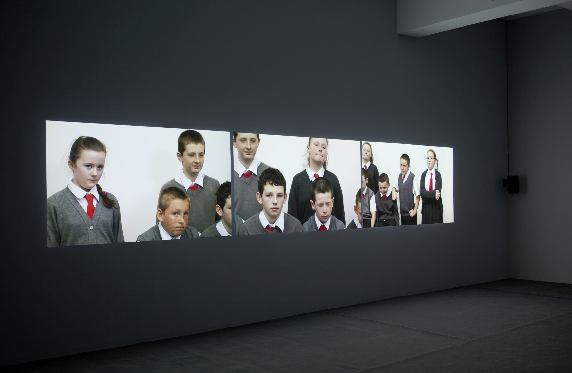 Three horizontal projections side by side in a dark room depict ten children in school uniforms with red ties and grey vests or sweaters against a white wall. 