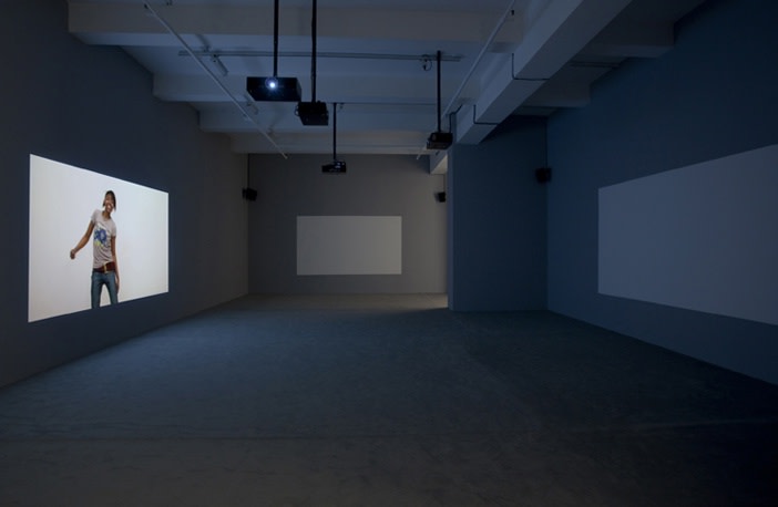 A dimly lit grey room with three projection screens has a video playing on the left wall portraying a girl dancing in a t-shirt and jeans. 