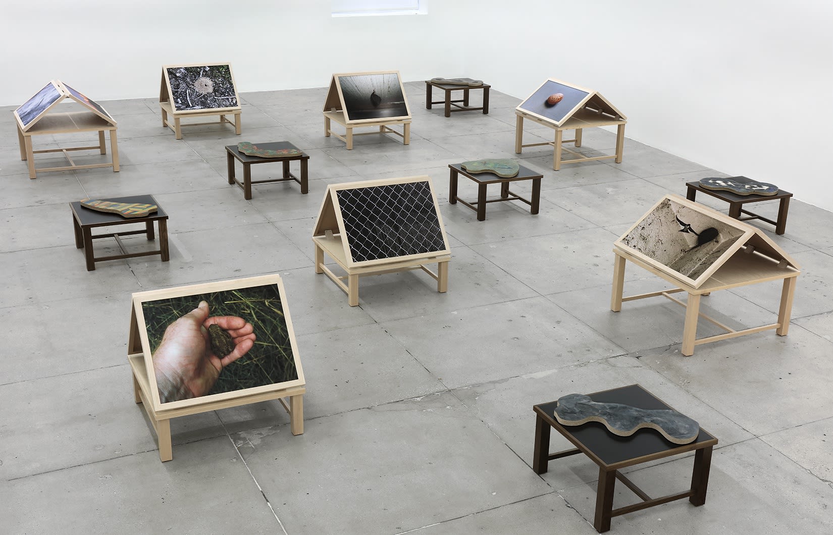 13 wooden platforms display color photographs and flat sculptures over a concrete floor. 