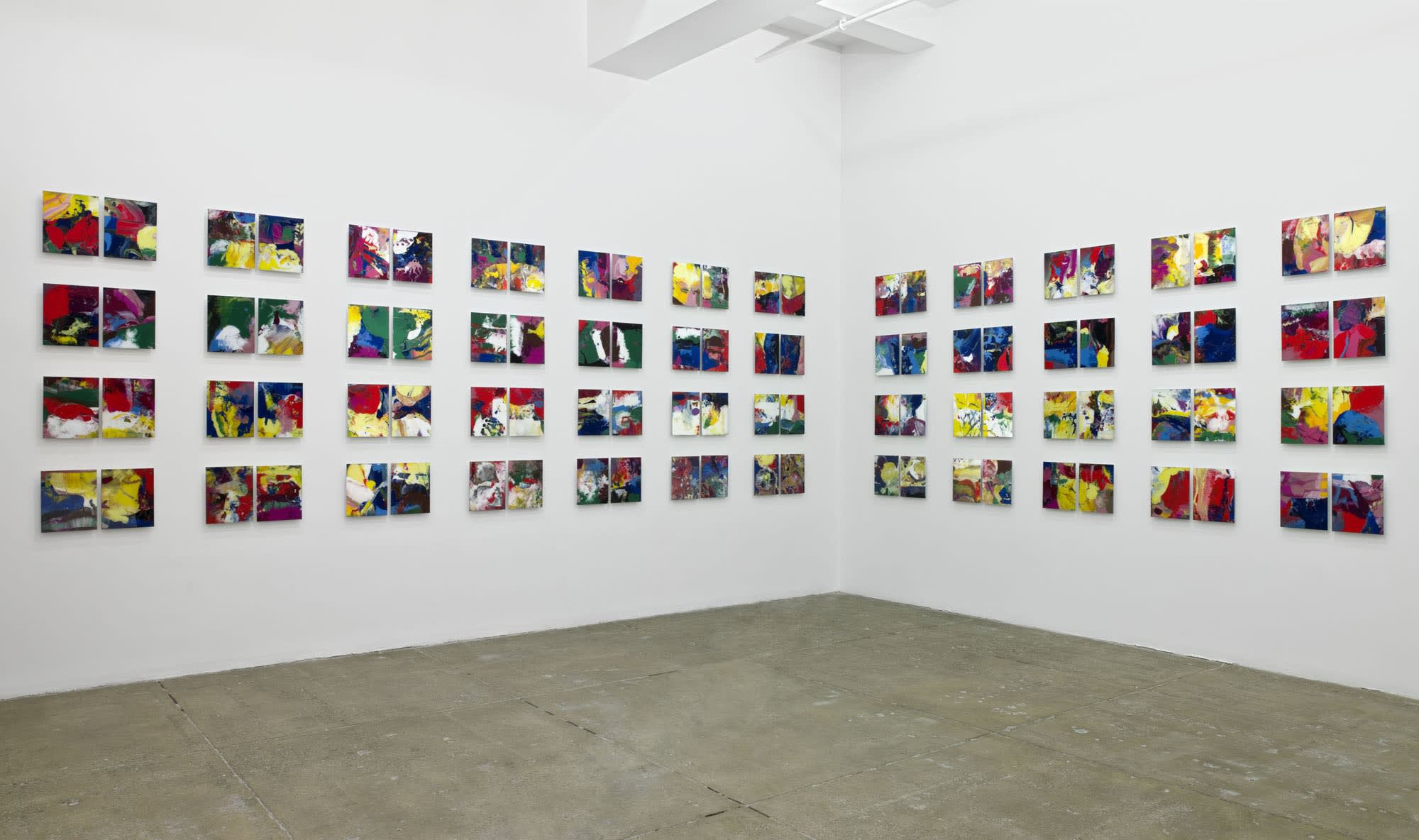 96 small, colorful paintings hang in rows of 4 and columns of 2 in a corner of a white room. 