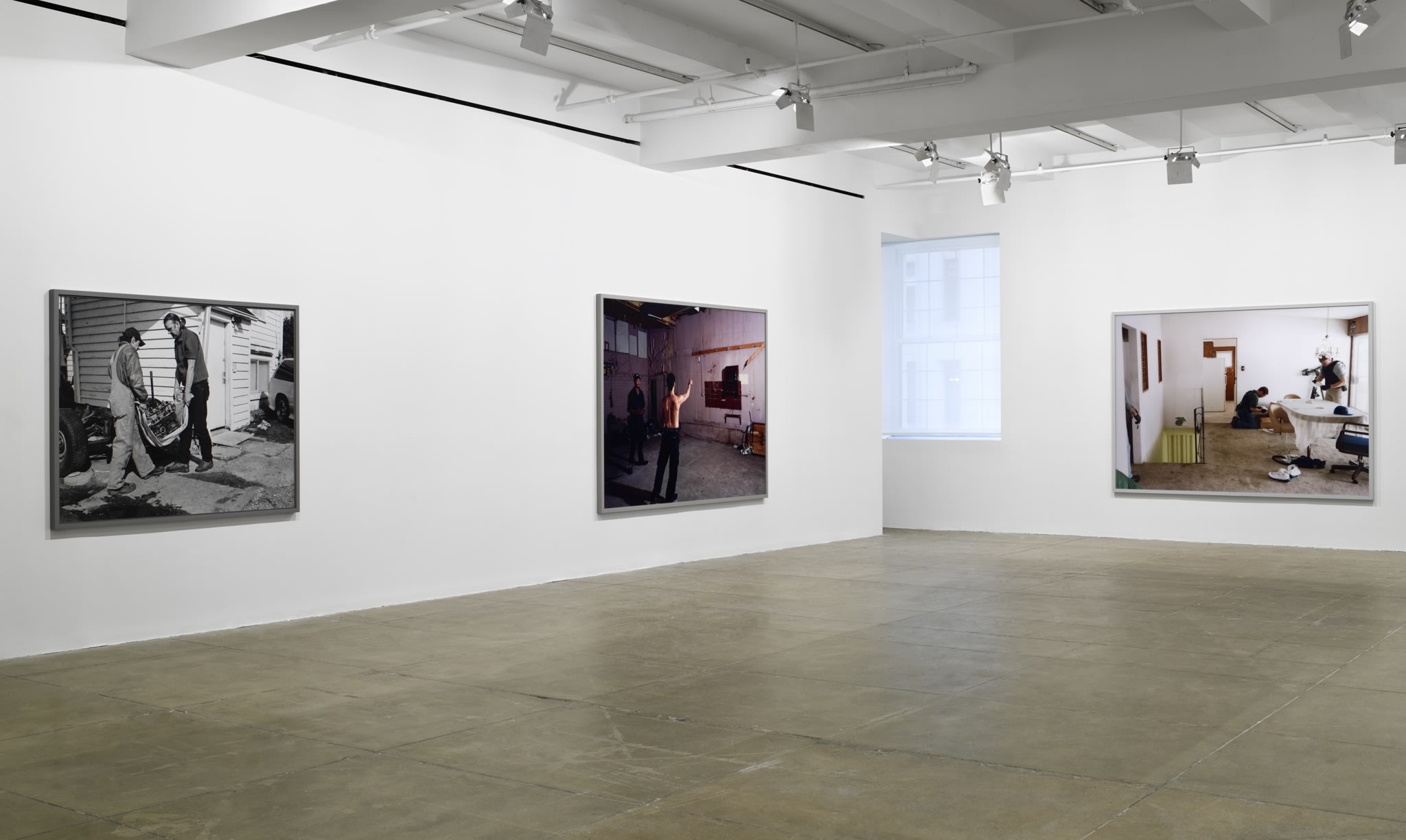 3 large, framed photographs of people involved in various activities hang in the corner of a white room. There is a window in the middle. 