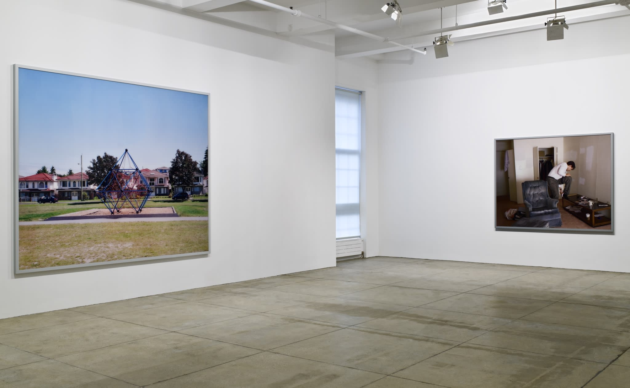 2 large, framed color photographs hang in the corner of a white room; on the left, a playground and on the right, a man in a dingy living room. There is a window in-between them. 