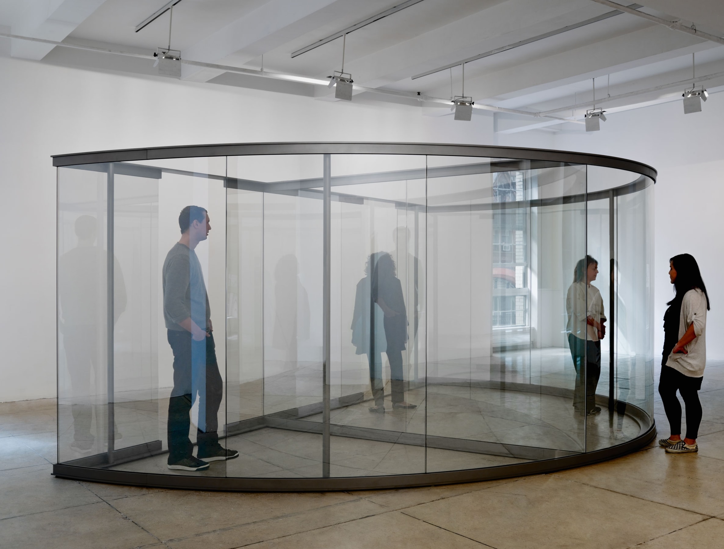 Various people move through a steel-framed half circle with glass panels, distorting their reflection. 