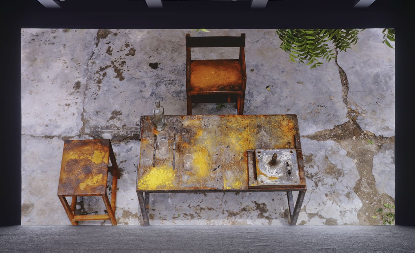 A large projection depicts a distressed wooden table and chair outdoors with two glasses and a bottle on top. 