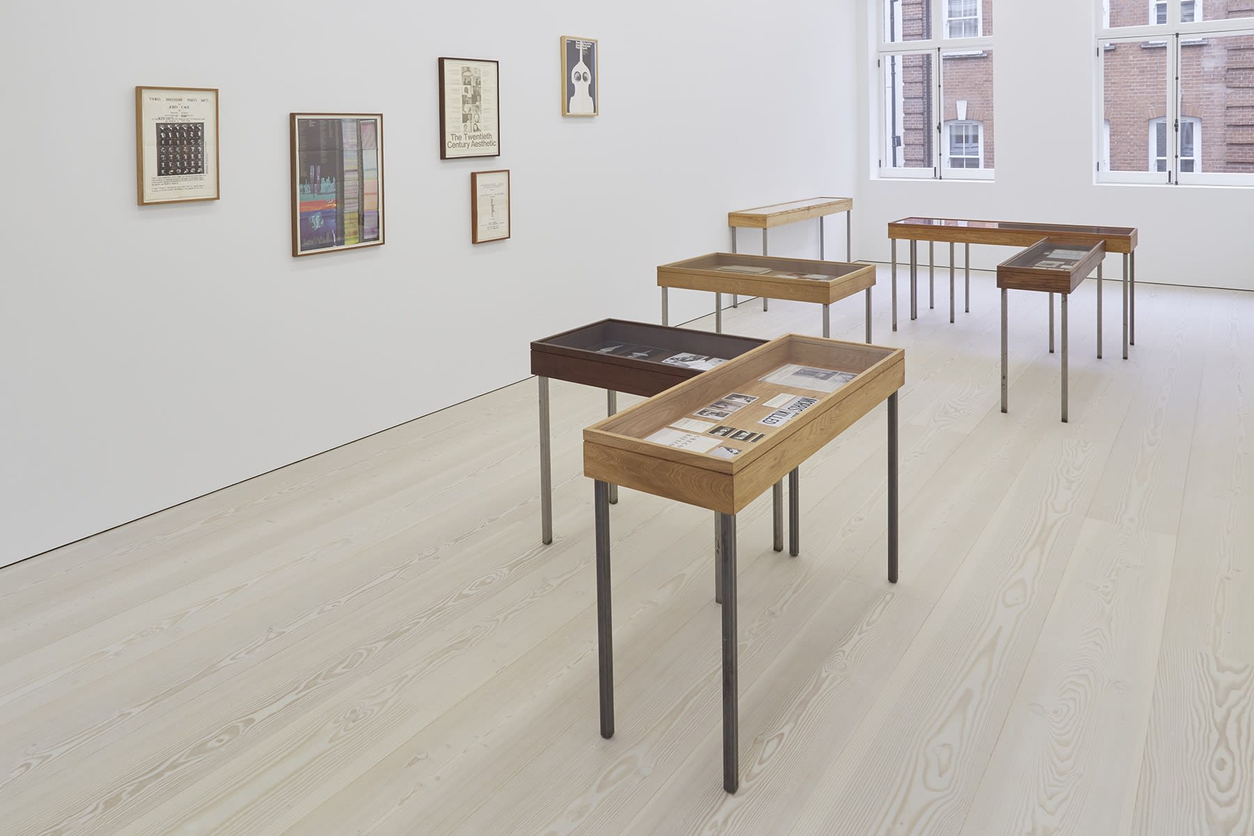 In a white space with light wood floors, several tables with glass tops act as display cases for various drawn works. Both colorful and black and white paintings and prints hang from the walls.