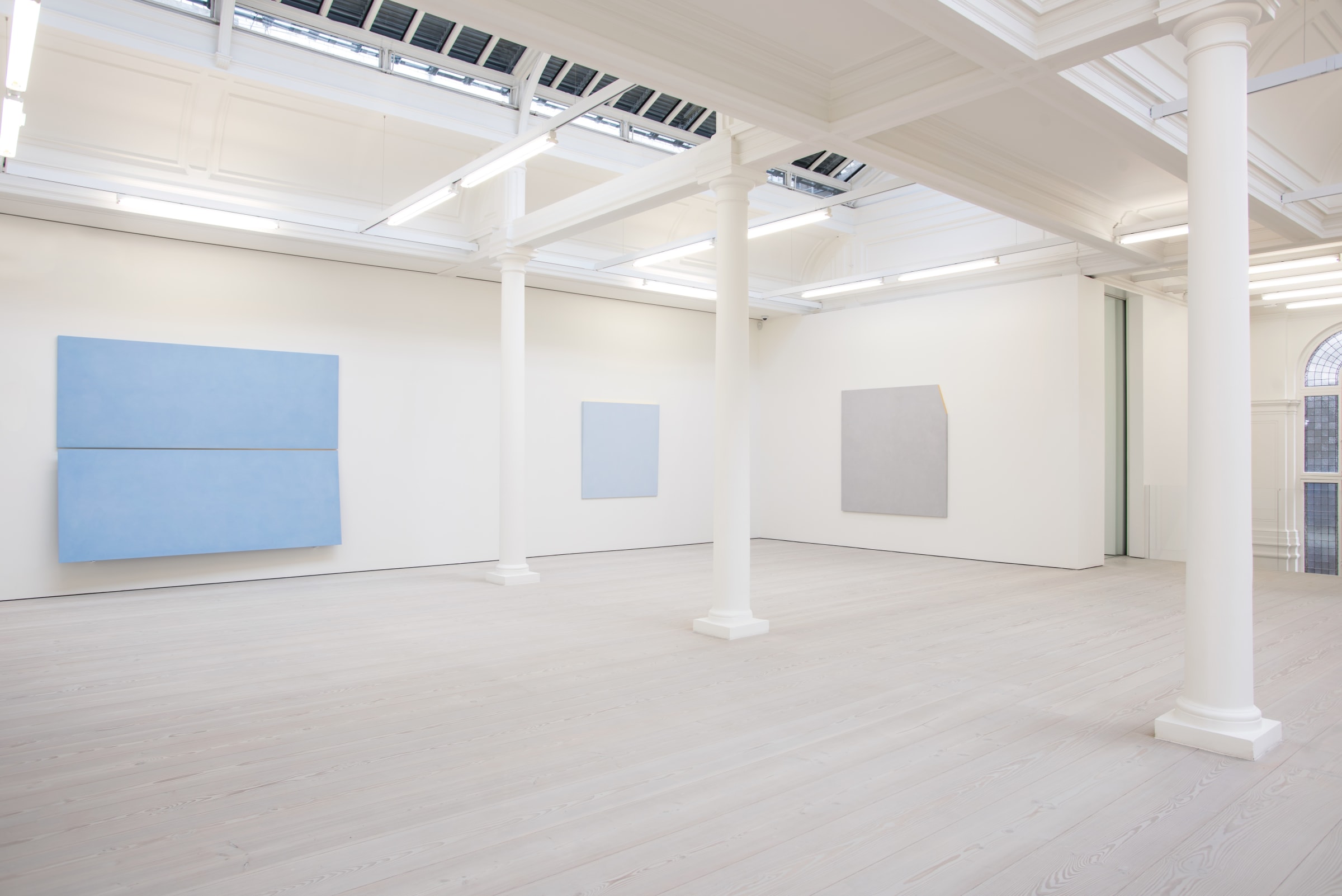 In a large white space with columns, two large light blue paintings hang, with one light grey painting.