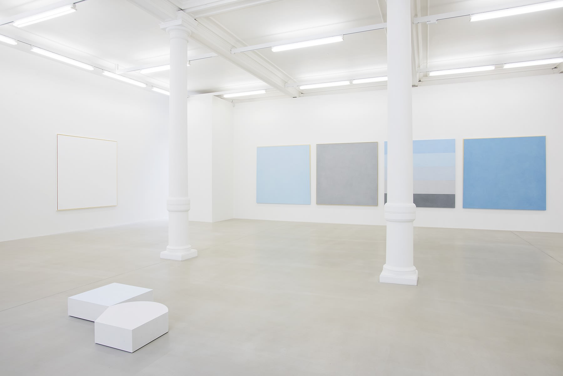 Various paintings of light shades (blue, grey, white) line the walls of an all white space, with short white sculptures on the floor.