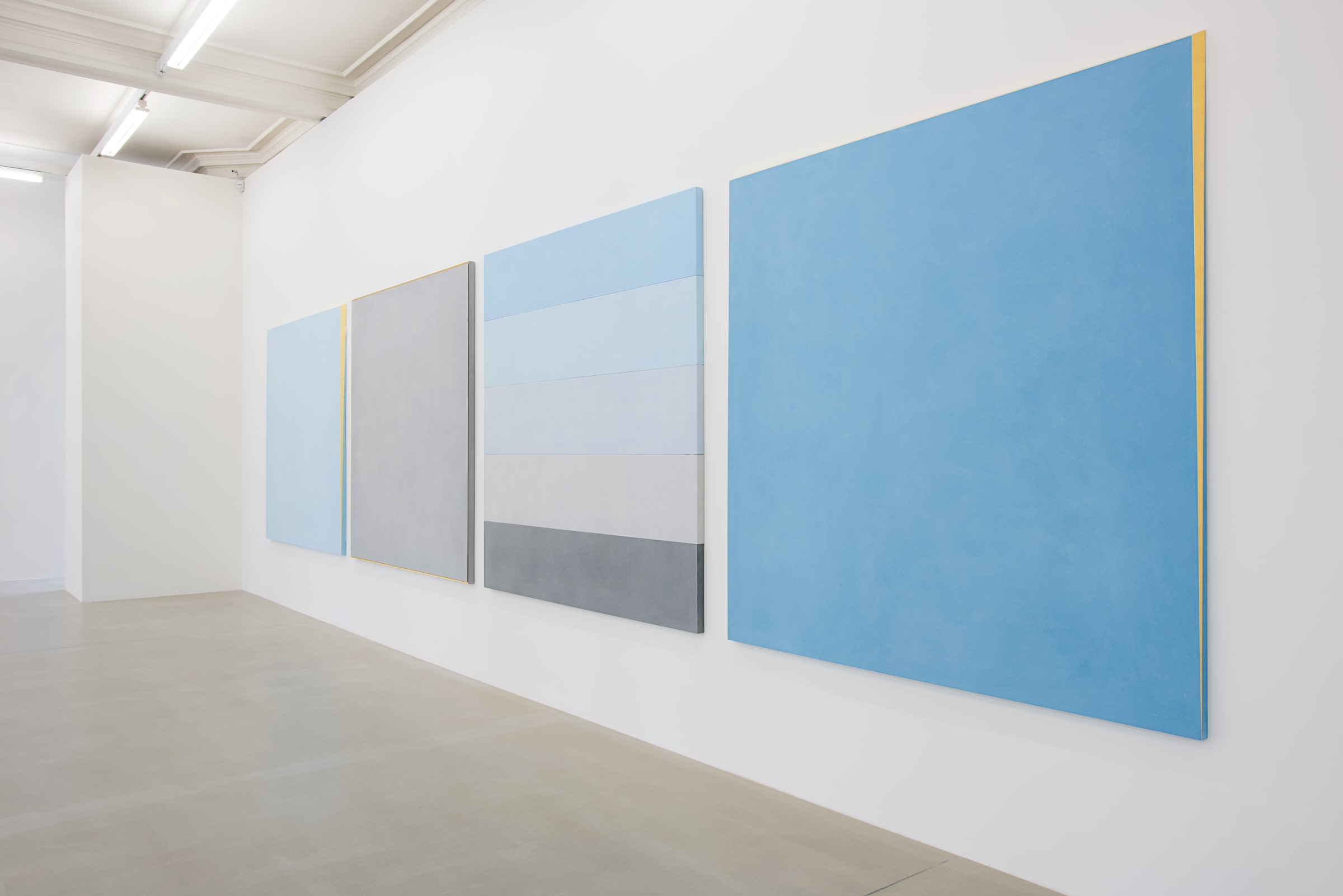 Four paintings hang on a white wall. From the left: solid light blue with gold framing, solid light gray with gold framing, 5 strips: light blue, lighter blue, light grey, grey,  darker grey, and finally, sky blue with gold framing.